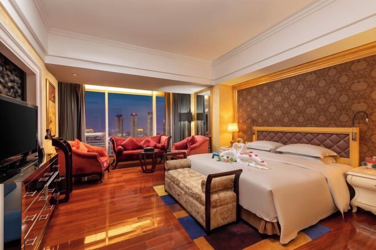 A hotel room with a large bed, a television, a sofa, and a view of the city at dusk.