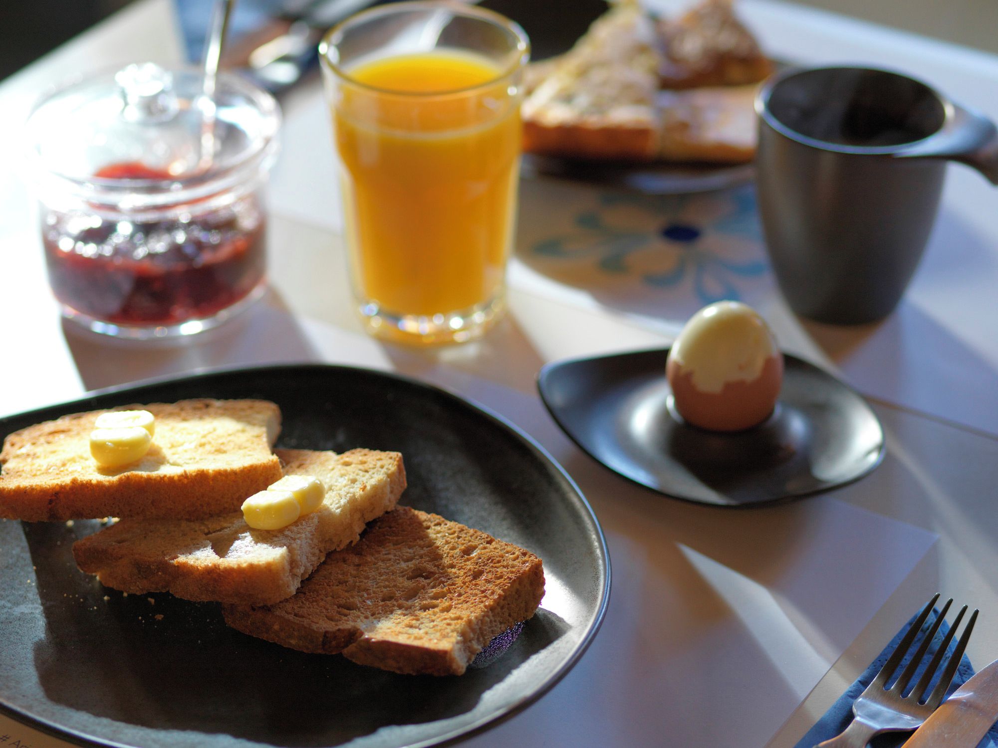 Une table mise pour le petit-déjeuner avec des tartines, du beurre, de la confiture, du jus d’orange et une tasse de café.