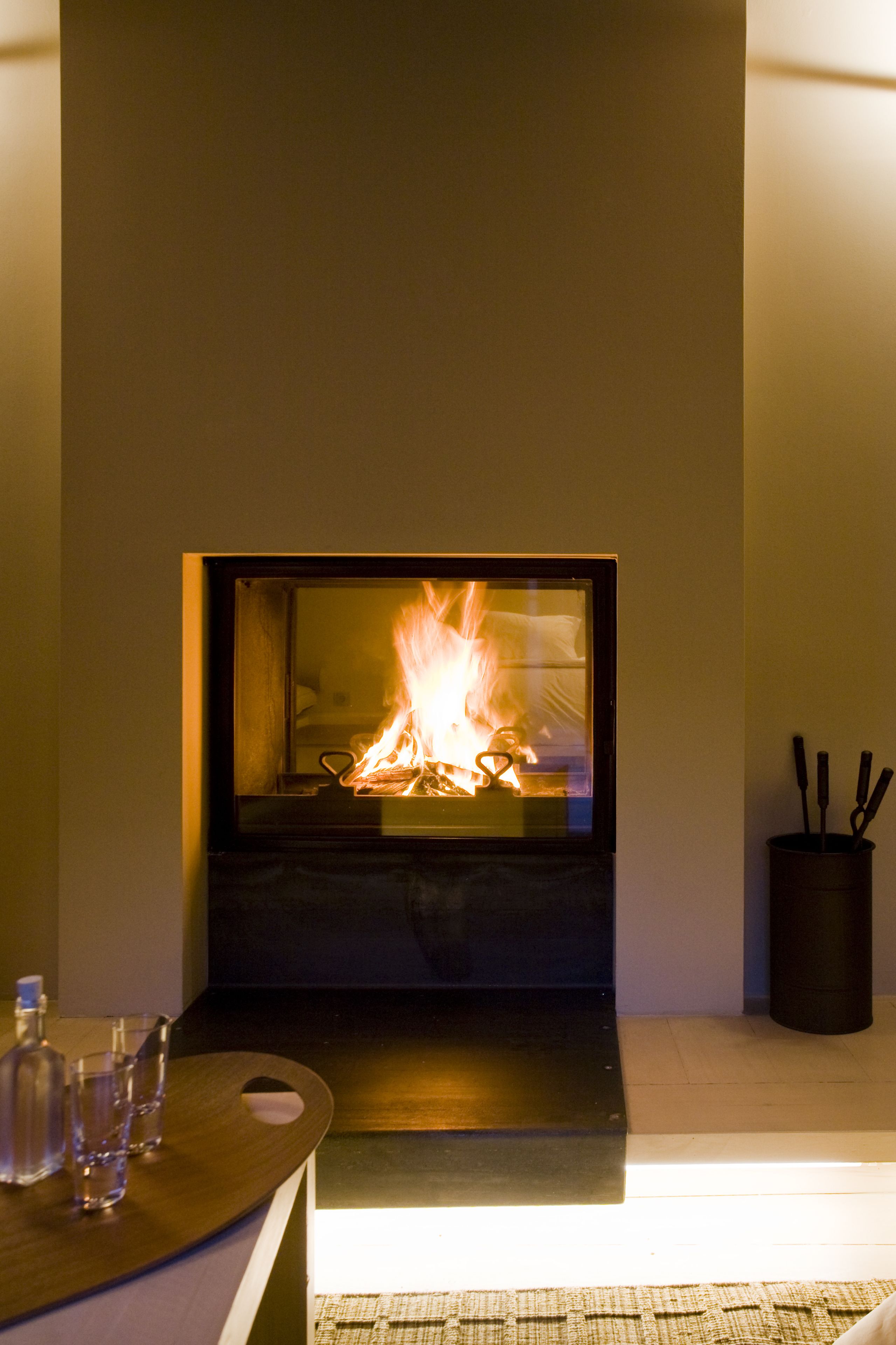 Una chimenea moderna con fuego visible, una mesa de madera con vasos y una botella, y un candelabro negro a la derecha.