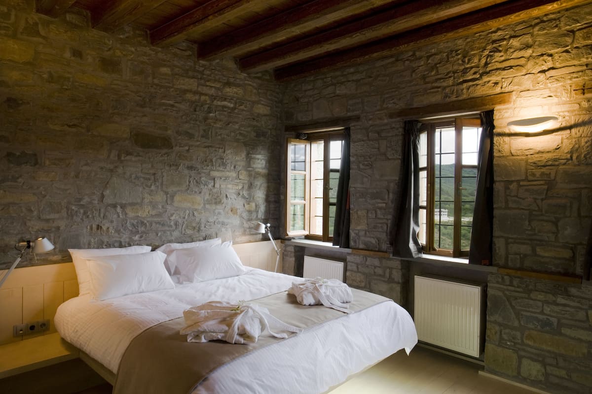 Una habitación con paredes de piedra, una cama grande con ropa blanca, una mesita de noche con lámpara y una ventana con cortinas.