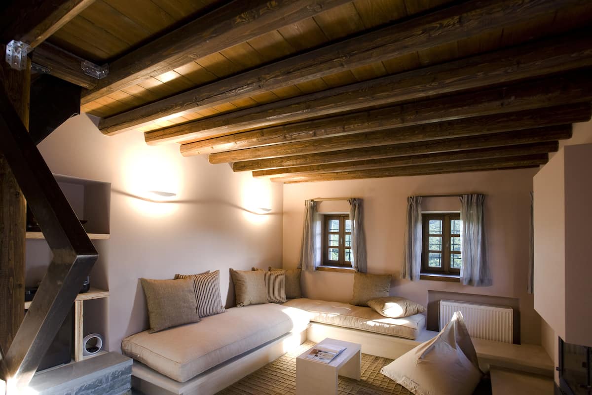 Un dormitorio acogedor en el ático con techo de madera, un sofá, dos ventanas con cortinas y un radiador.