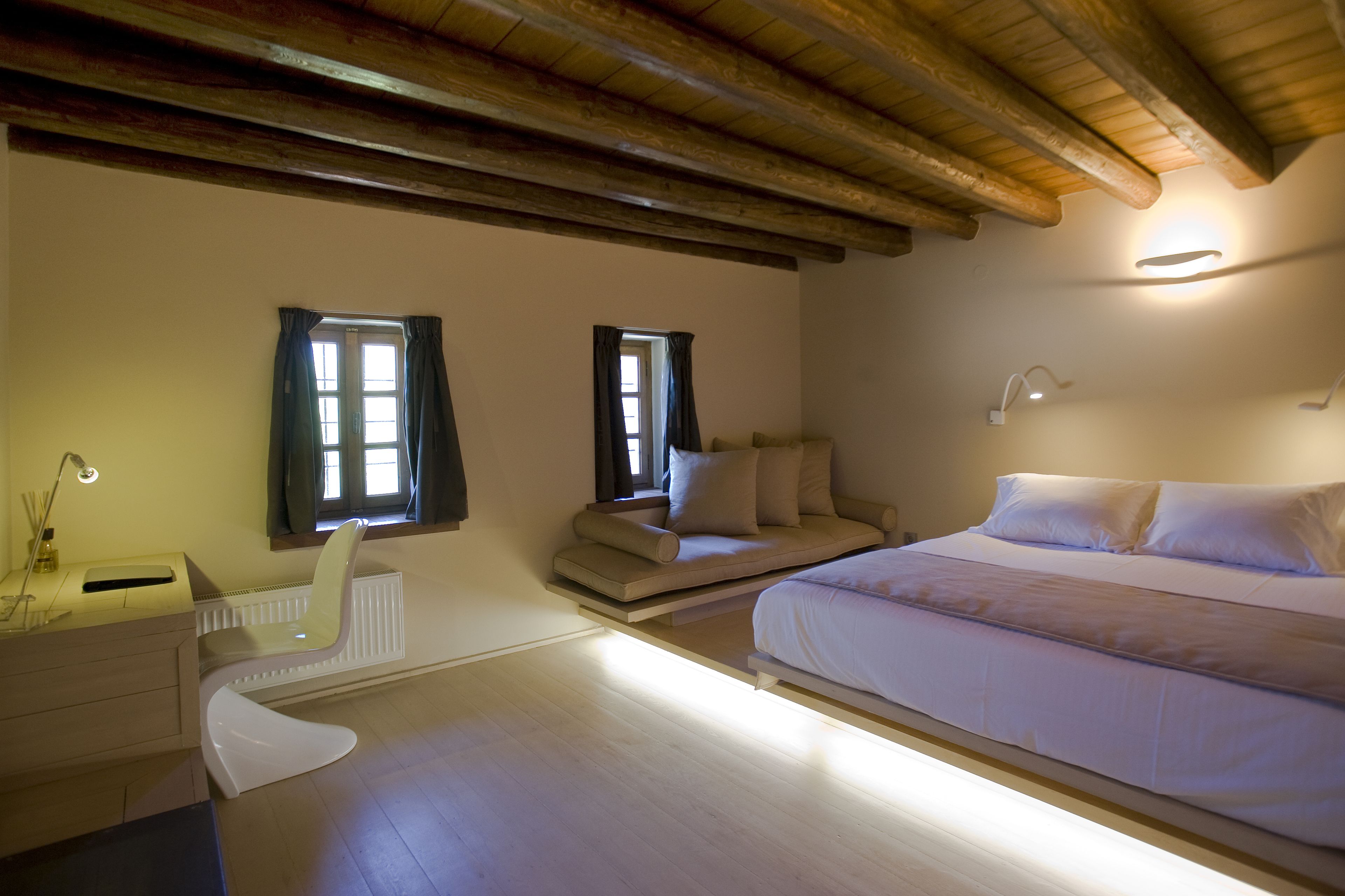 A bedroom with a bed, a desk, and a chair. There are two windows with curtains.