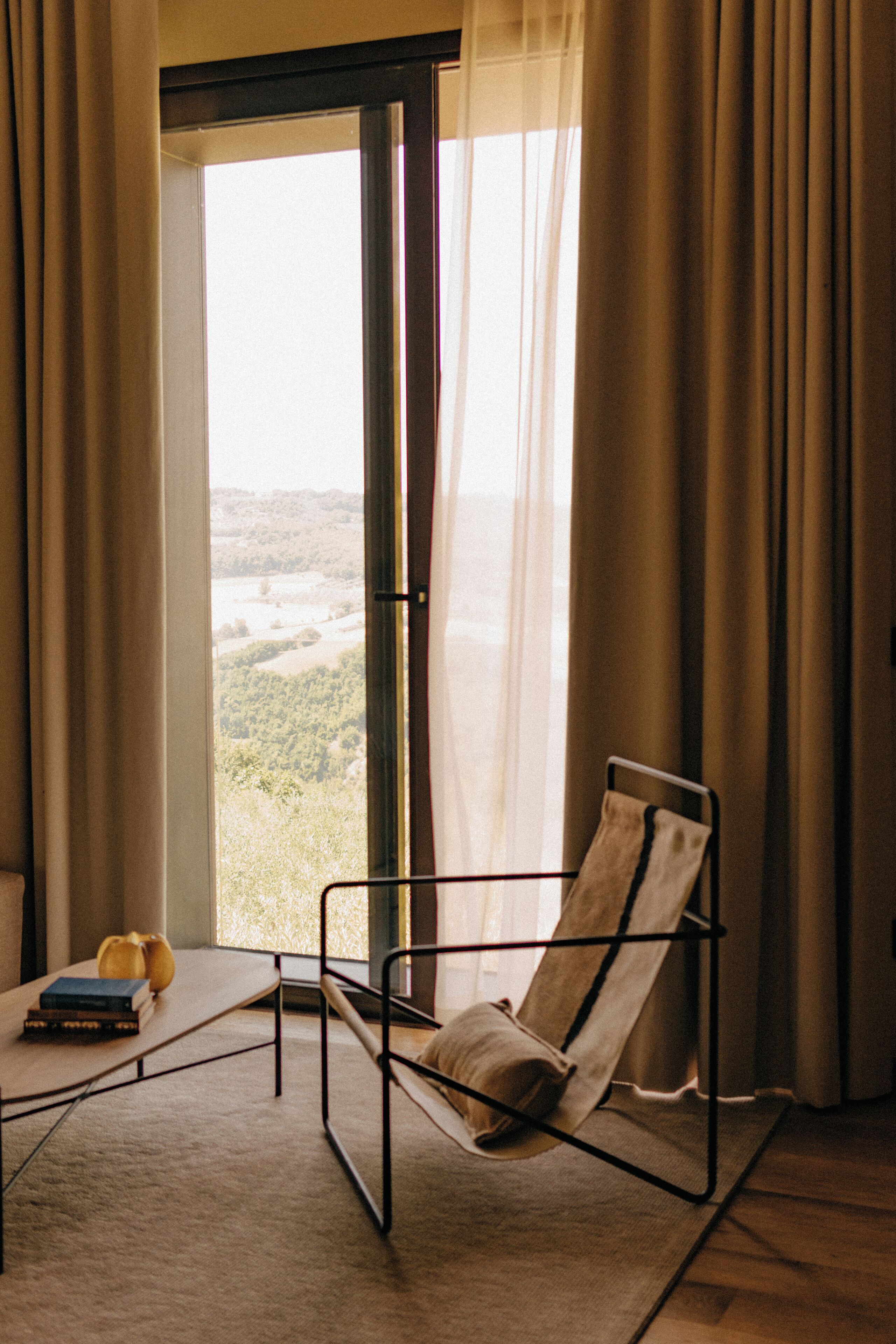 A room with a view of a landscape through a large window, a chair, a small table with books, and a pillow on the chair.
