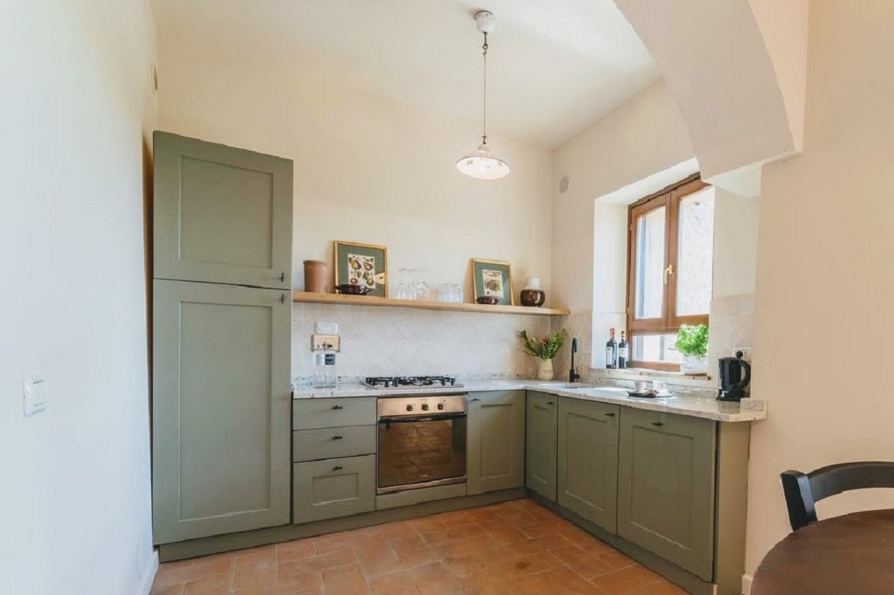 Cocina compacta con estufa a gas, horno y armarios verdes. Cuenta con una ventana que proporciona luz natural y una lámpara colgante.