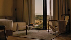 Une chambre d’hôtel moderne dotée d’une grande fenêtre, d’un canapé, d’un fauteuil, d’une petite table avec des livres et offrant une vue sur la campagne.