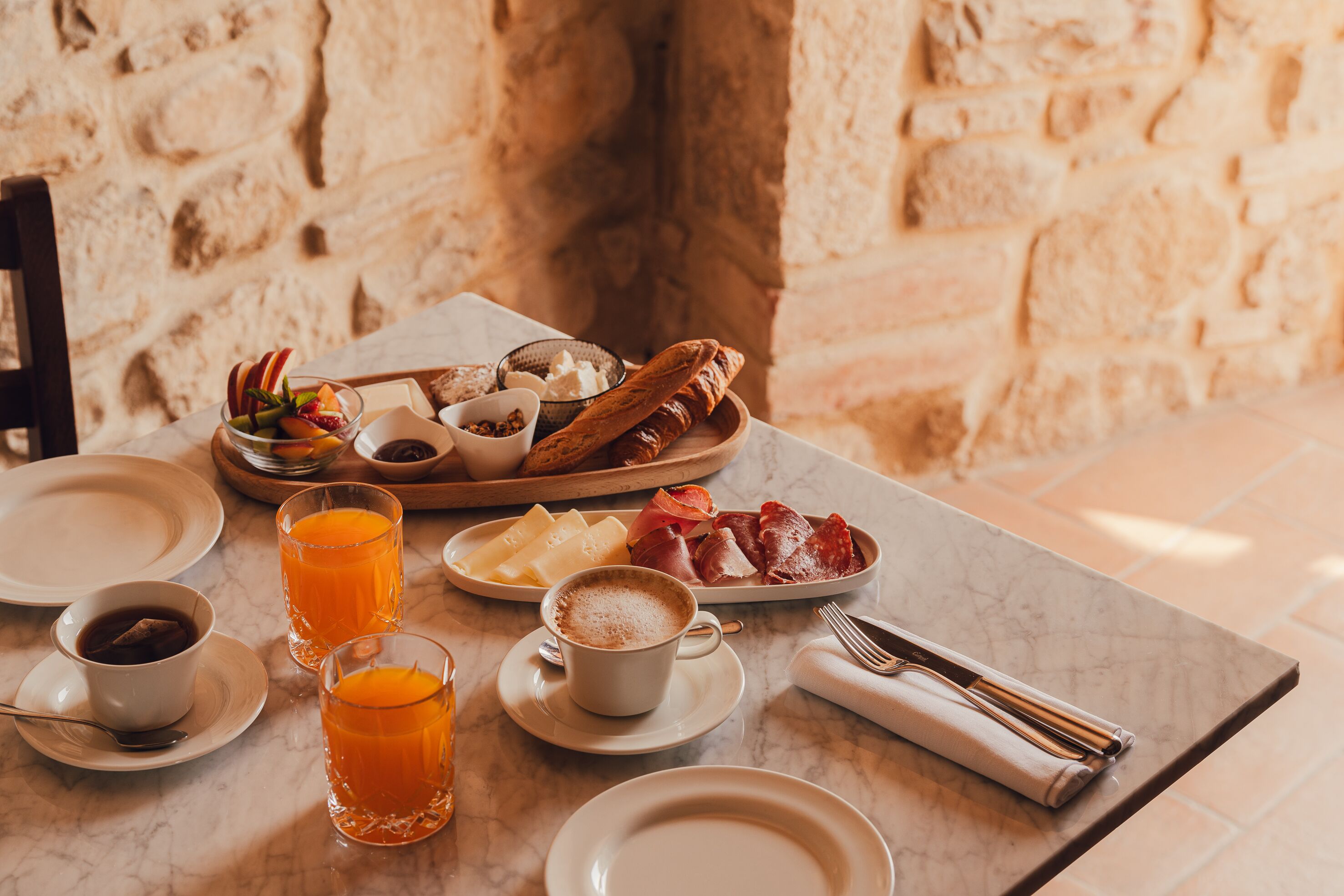A breakfast setting with a stone wall background, featuring a variety of foods and drinks on a marble table.