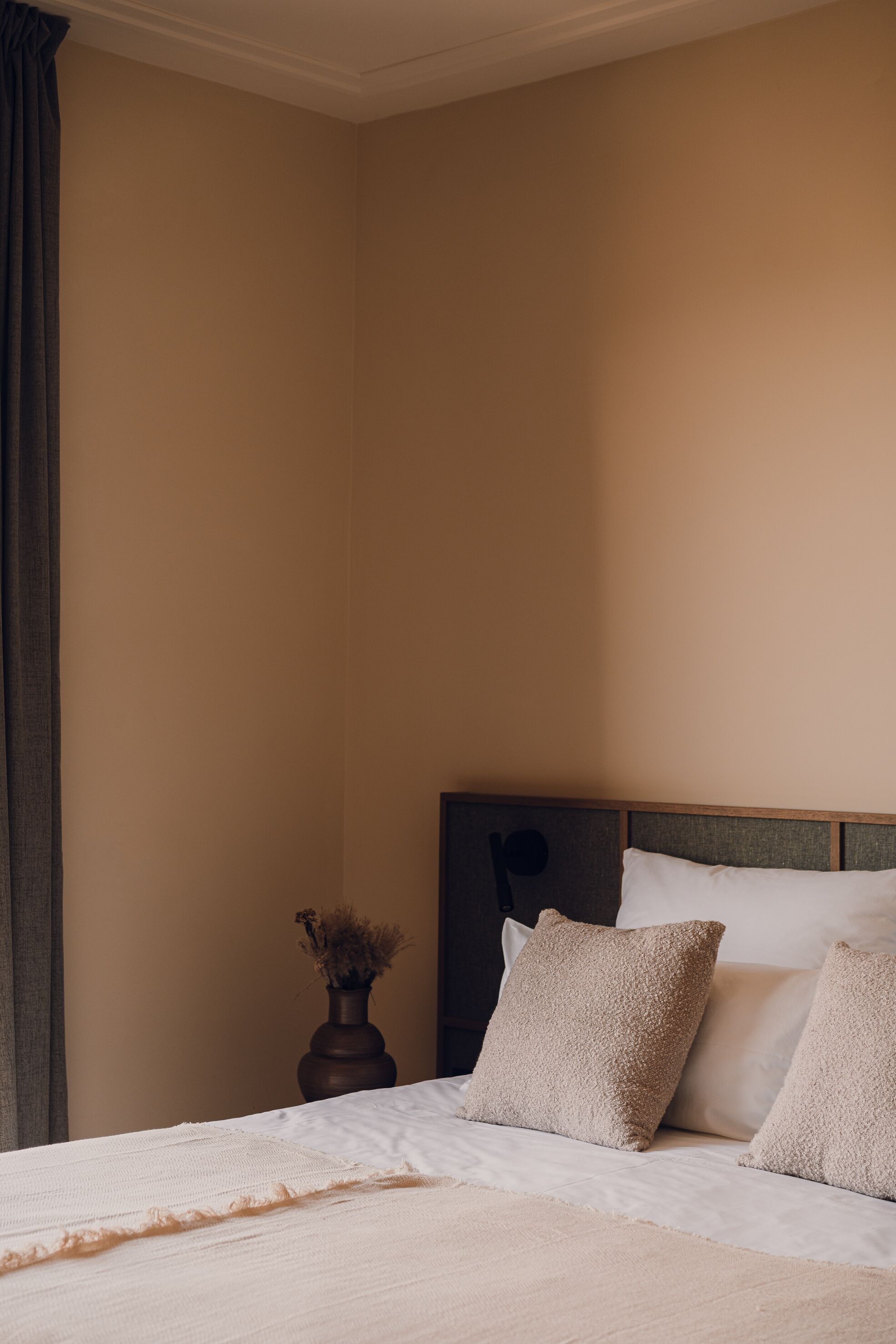 A neatly made bed with white linens and two beige pillows, a dark wooden nightstand with a vase of dried flowers, and a beige wall.