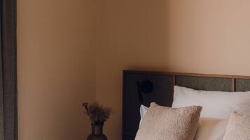 A neatly made bed with white linens and two beige pillows, a dark wooden nightstand with a vase of dried flowers, and a beige wall.