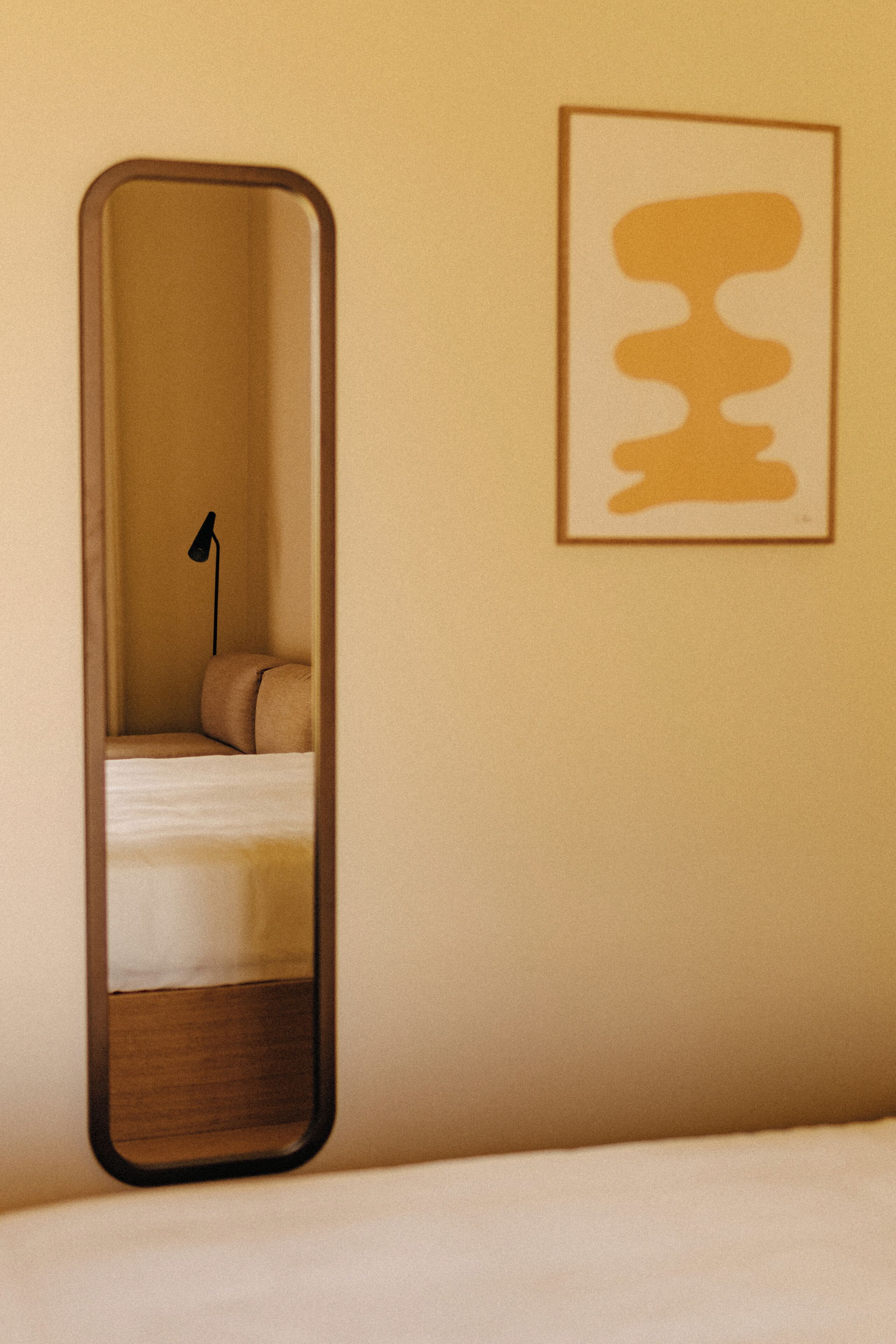 Een slaapkamer met een bed, een bedlamp, een spiegel en een abstract kunstwerk in een lijst aan de muur.