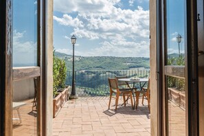 Un patio abierto con una mesa y sillas, una lámpara y vistas a un paisaje verde a través de una baranda metálica.