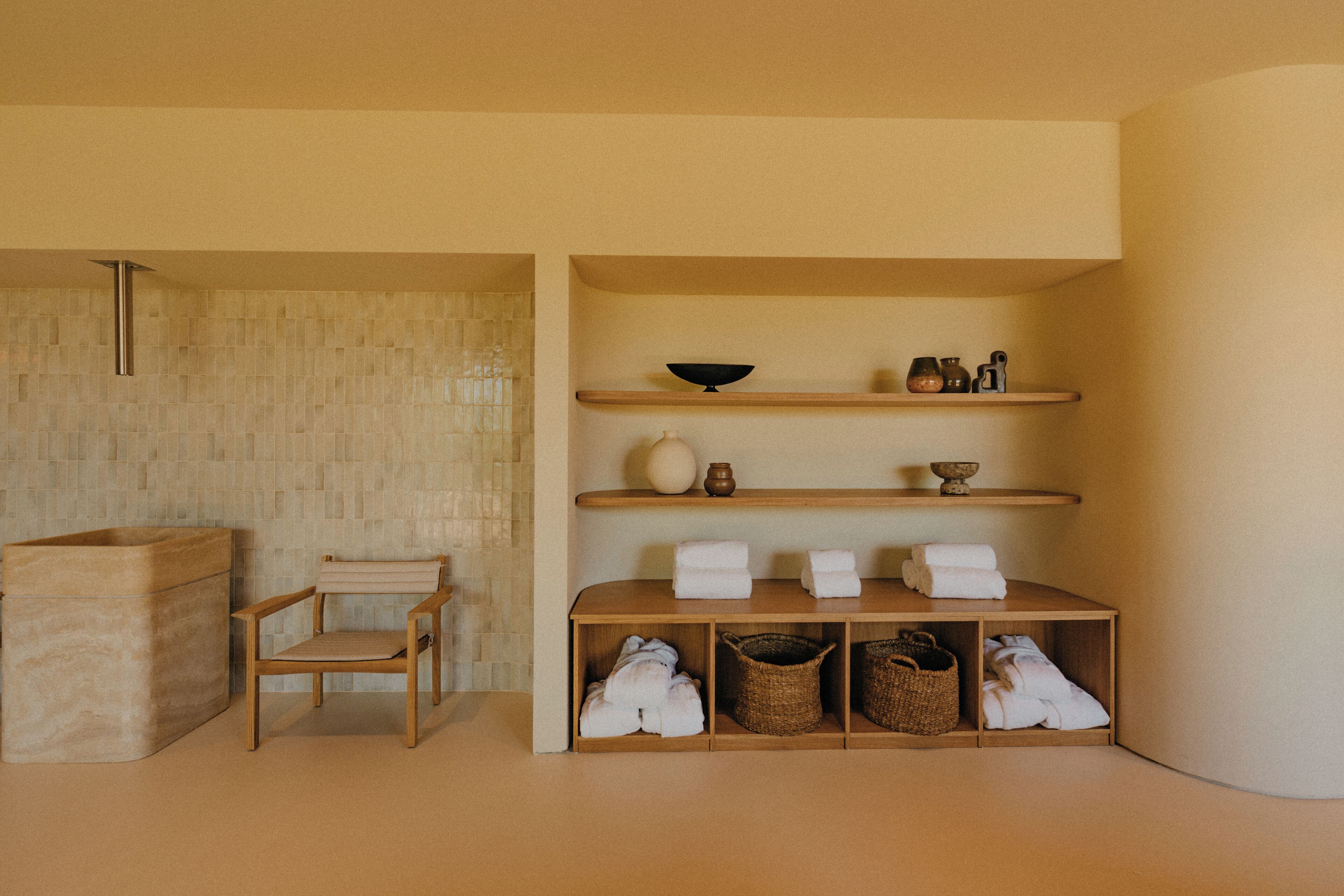 A minimalist bathroom with a wooden chair, shelves with towels, and a large basin.