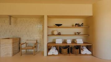 A minimalist bathroom with a wooden chair, shelves with towels, and a large basin.