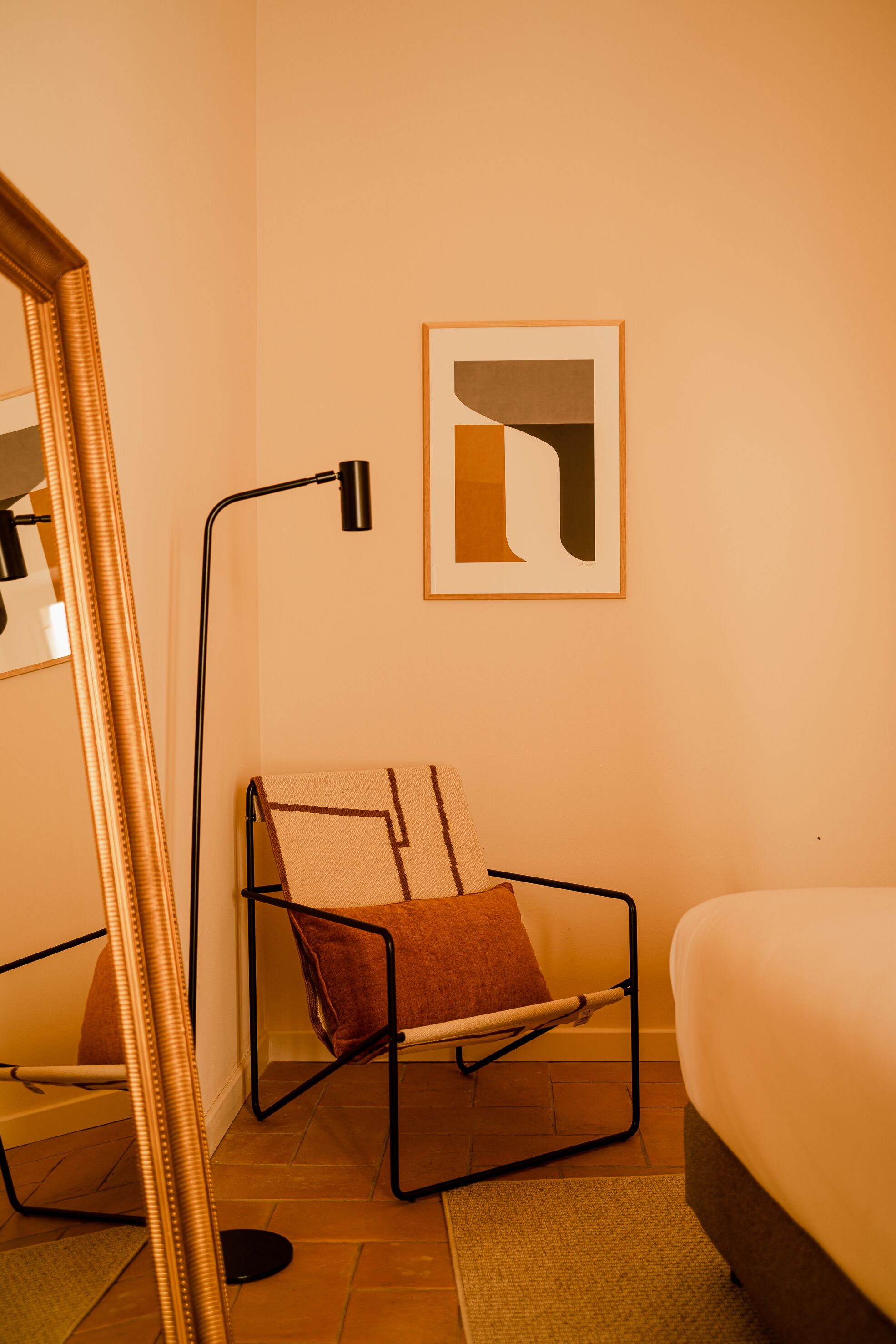 A room with a bed, a chair with a blanket, a floor lamp, and a framed abstract artwork on the wall.