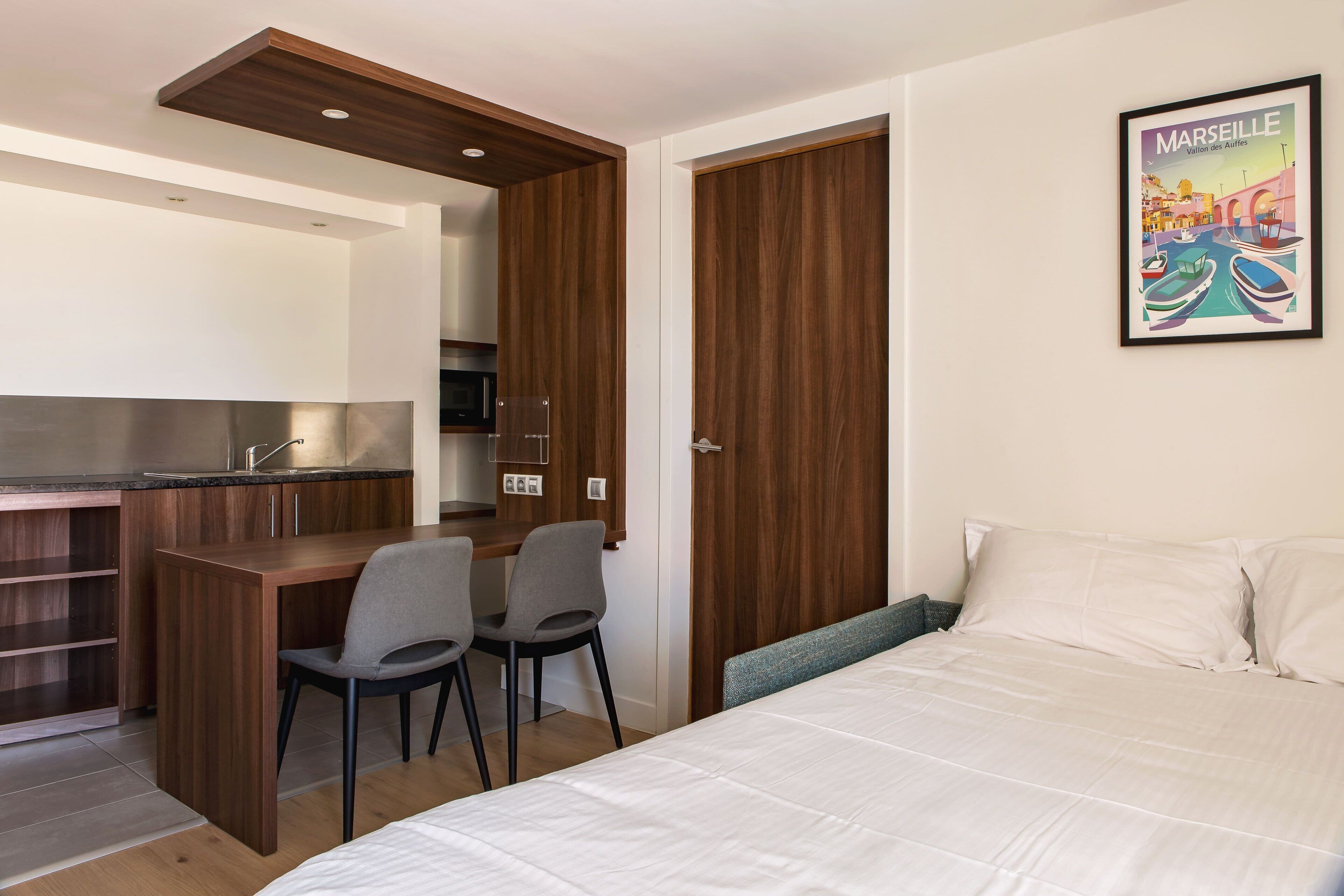 A bedroom with a bed, a desk, two chairs, a wardrobe, and a framed picture on the wall.