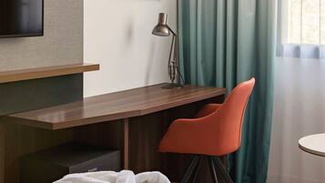 A hotel room with a bed, a desk with a chair, a TV mounted on the wall, and a window with curtains.
