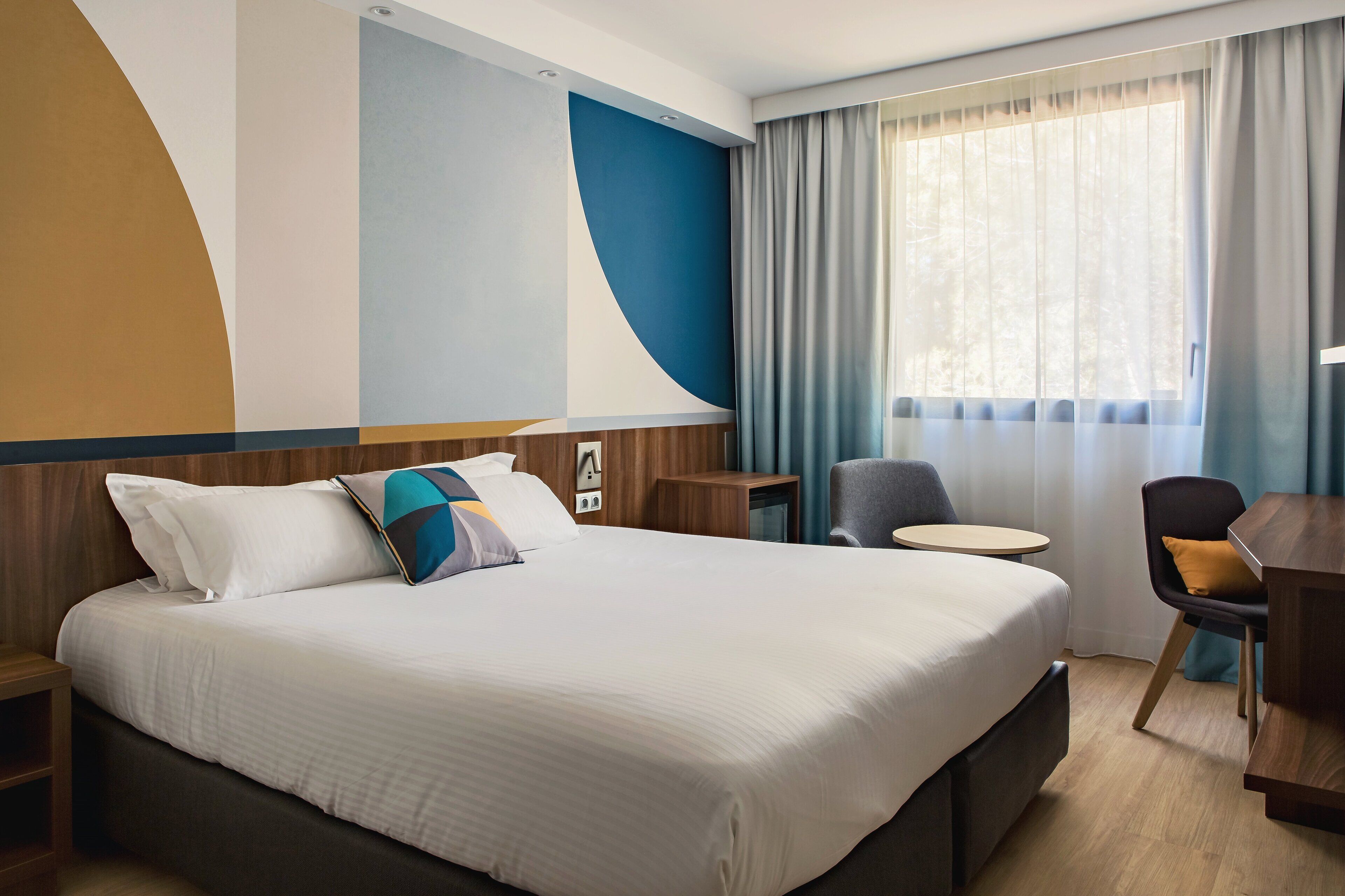 Una habitación de hotel con una cama grande, un escritorio, una silla y un ventanal con cortinas.