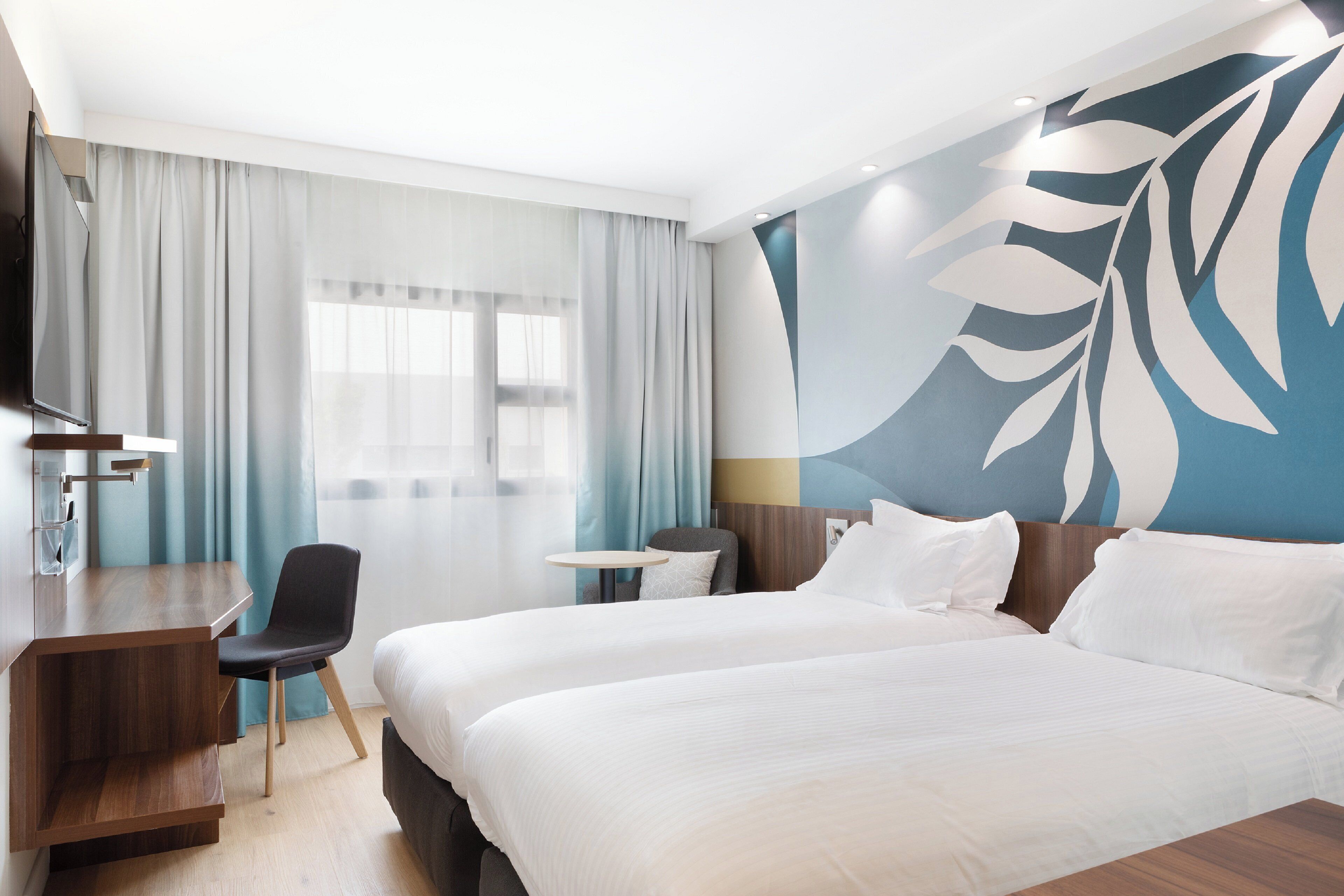 Een hotelkamer met twee bedden, een bureau, een stoel, een televisie en een groot raam met gordijnen.
