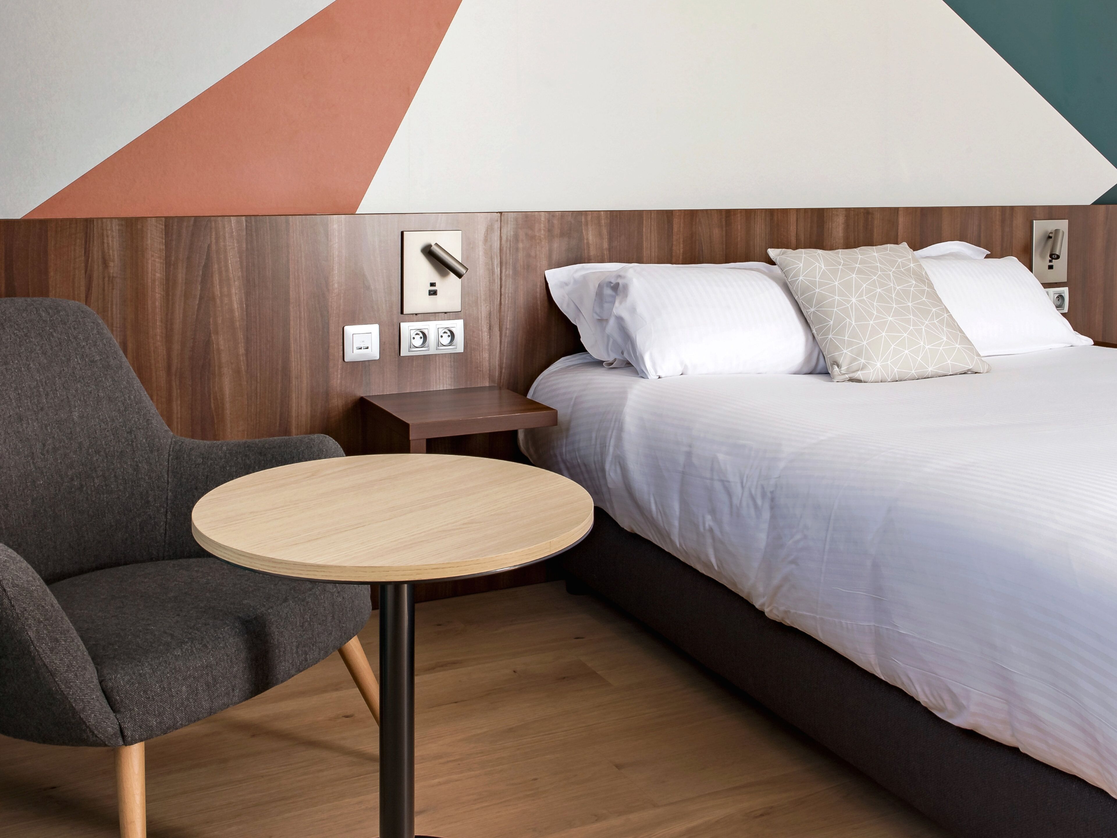 A hotel room with a bed, a chair, a small table, and a wall with a geometric design.