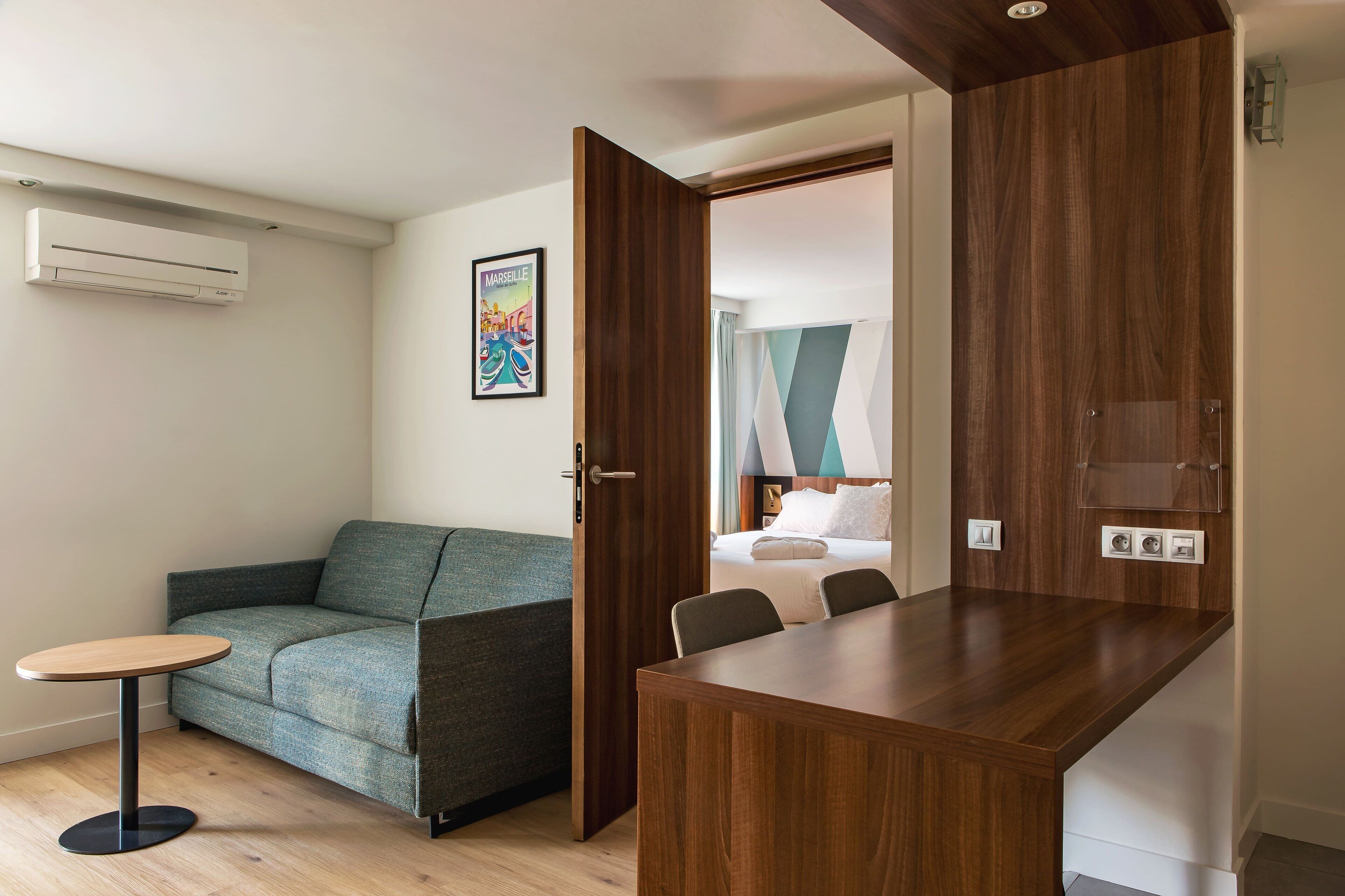A modern hotel room with a wooden dining table, a green sofa, a small round table, and a bed with a white headboard.