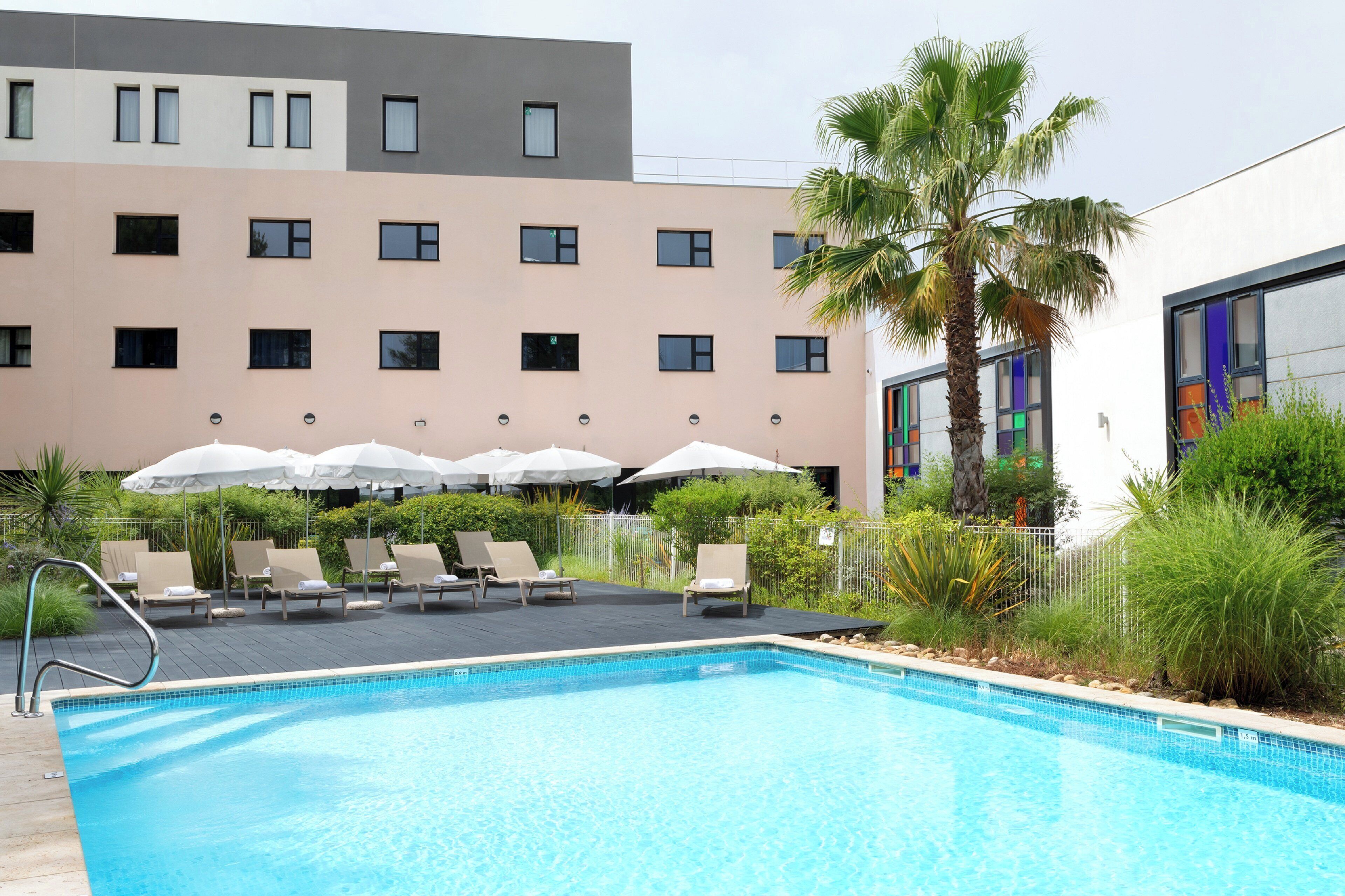 A swimming pool with lounge chairs and umbrellas, surrounded by greenery and a building with multiple windows.