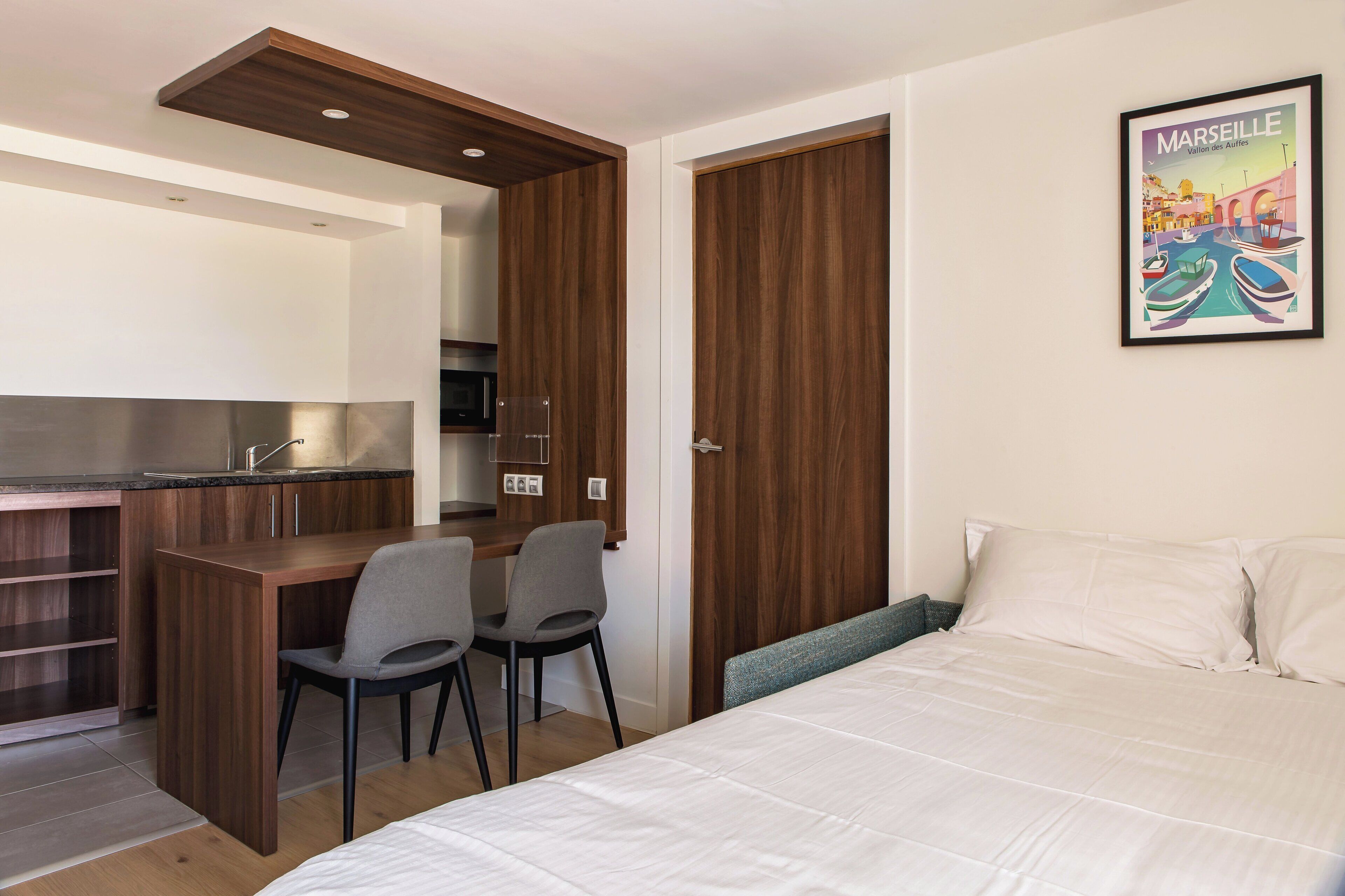 Een kleine kamer met een bed, een bureau, twee stoelen, een kitchenette en een deur.