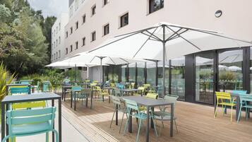 Een terras met tafels en stoelen, parasols, en een gebouw op de achtergrond.