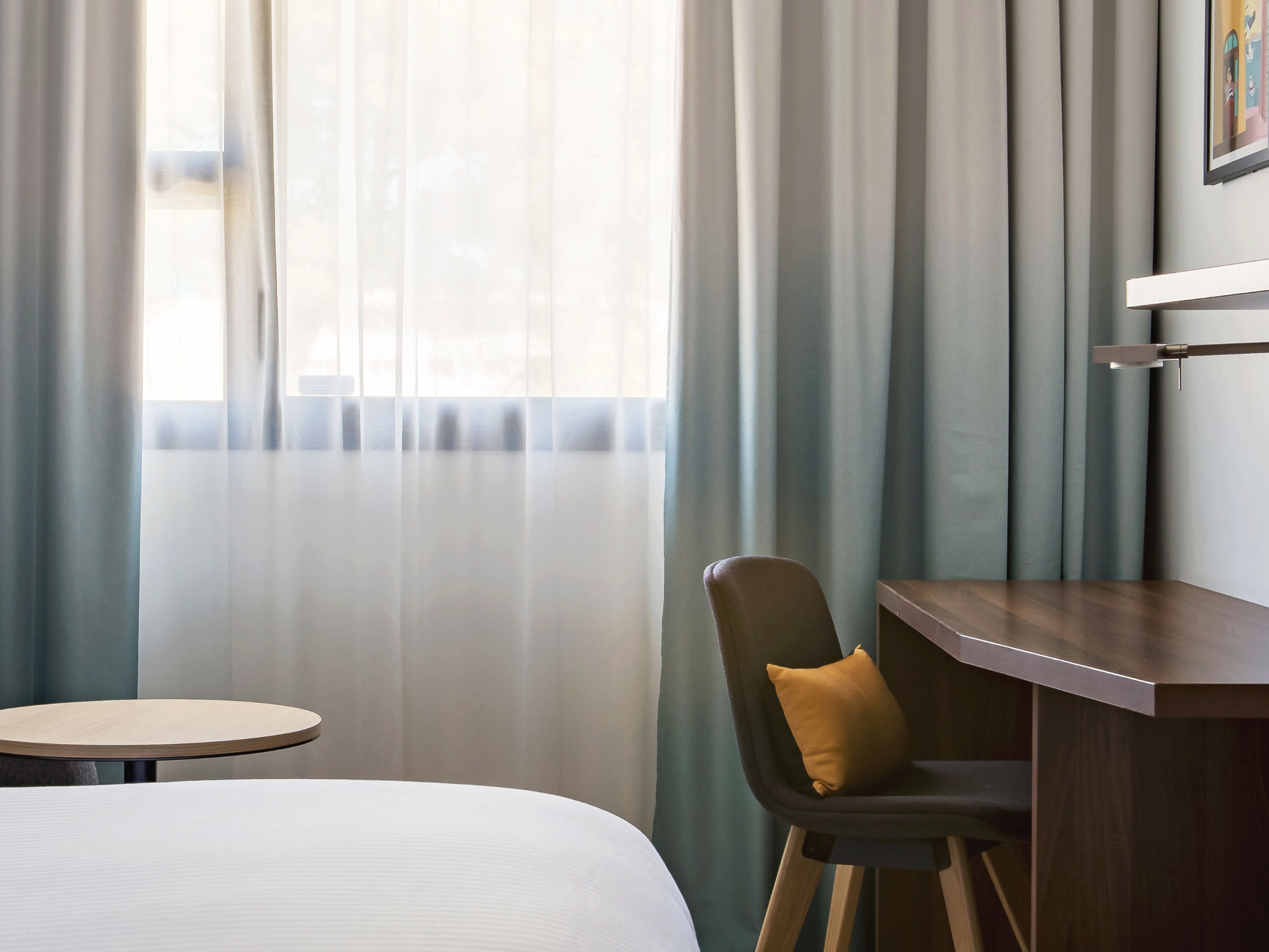 Et hotelværelse med en seng, et rundt bord, en stol med pude og et skrivebord med tilhørende stol.
