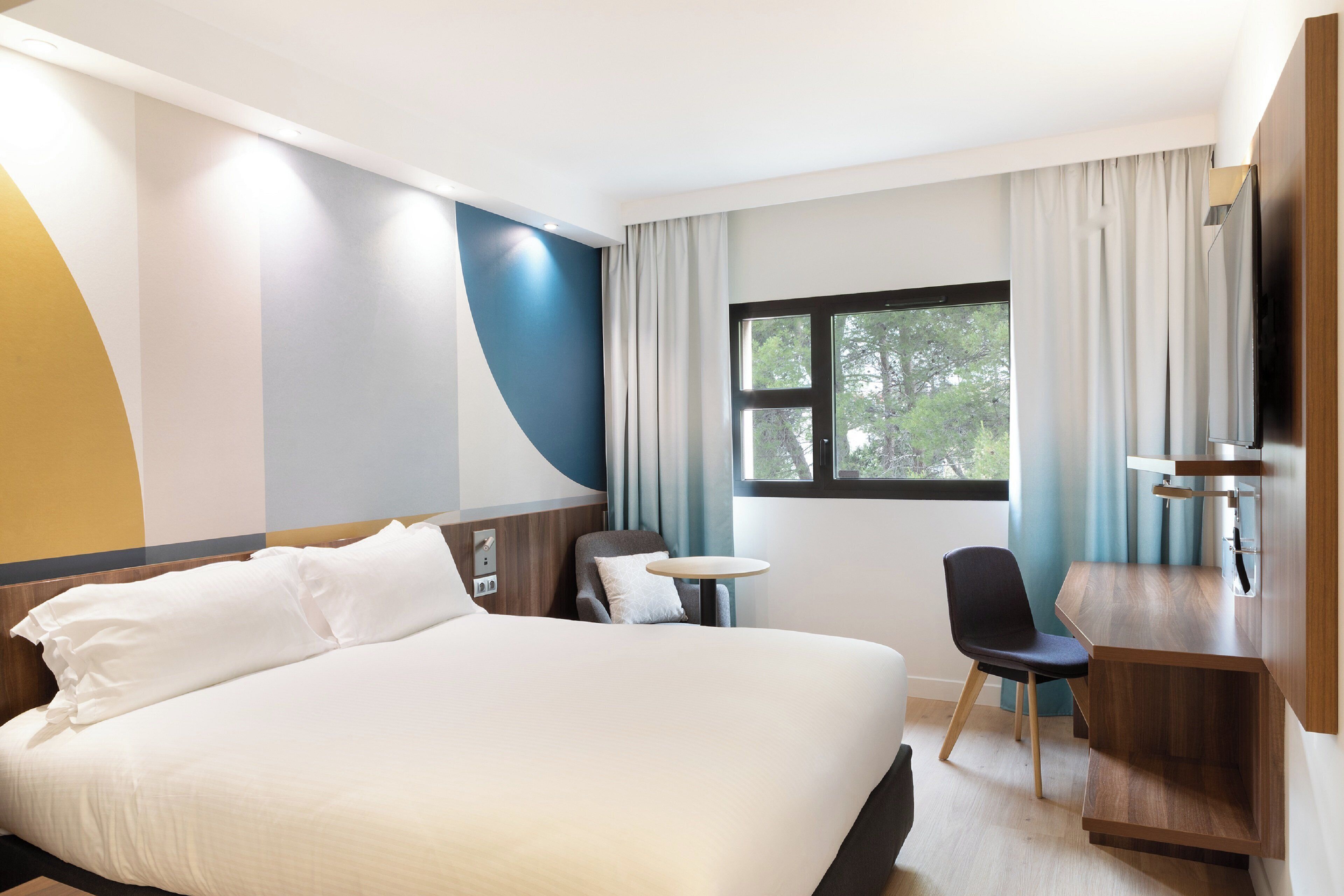 A hotel room with a large bed, a desk, a chair, and a window with a view of trees.
