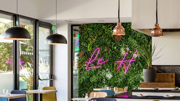 A modern restaurant interior with wooden tables and chairs, a green wall with a neon sign, and pendant lighting. - Holiday Inn Marseille Airport by IHG (Vitrolles)