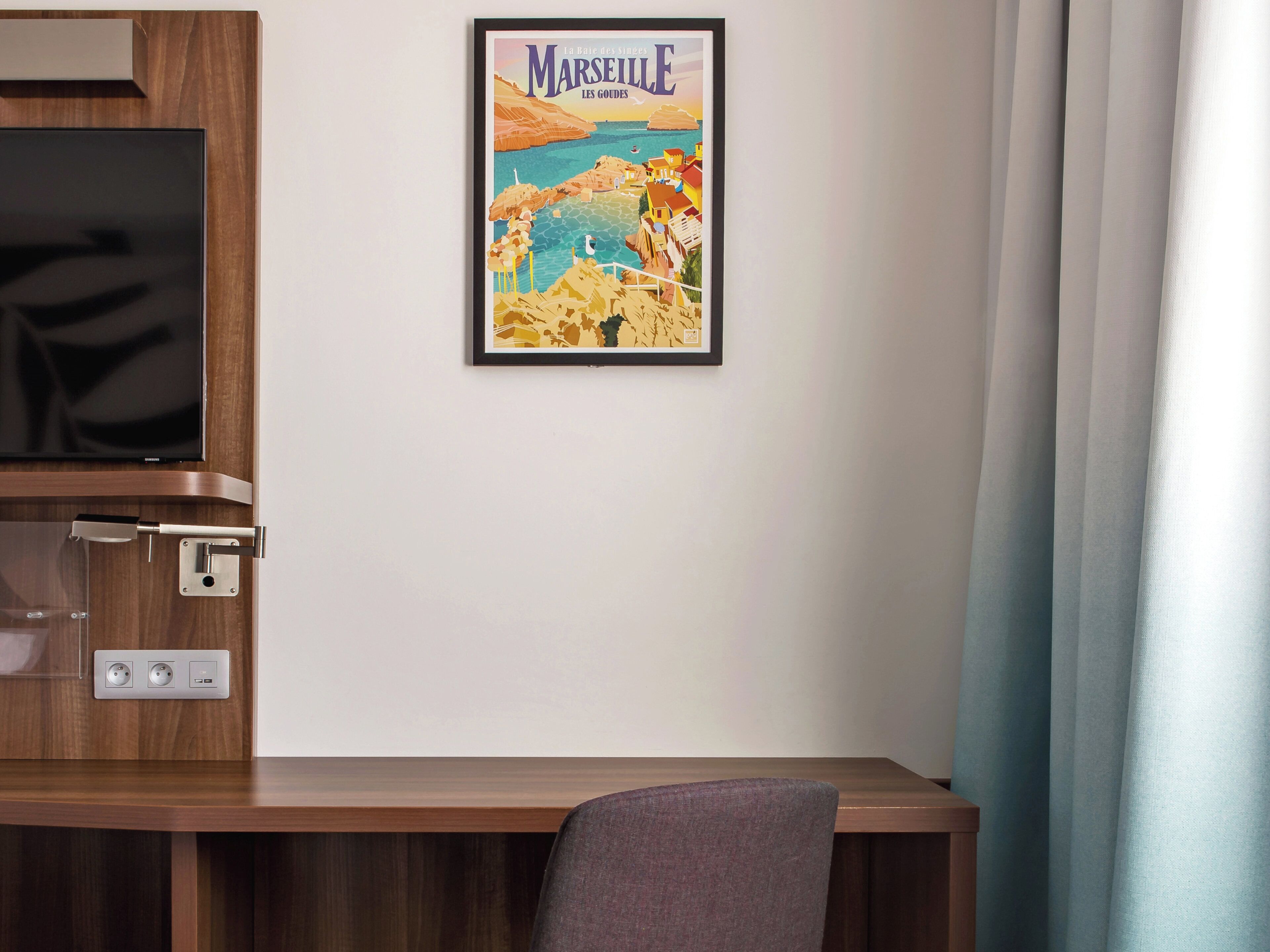 A hotel room with a wooden desk, a television, and a poster of Marseille.