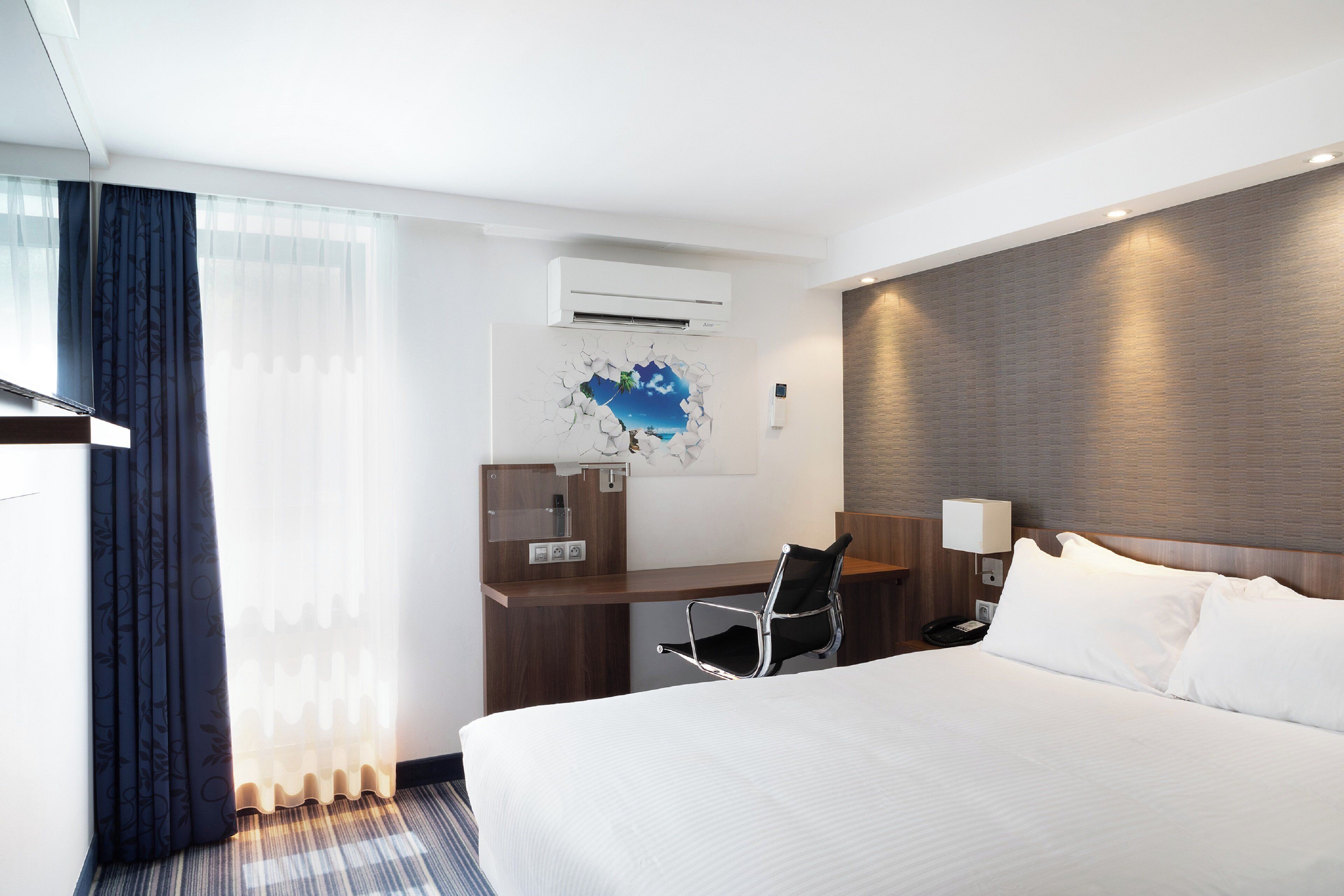 A hotel room with a bed, a desk, a chair, an air conditioning unit, and a wall-mounted lamp.