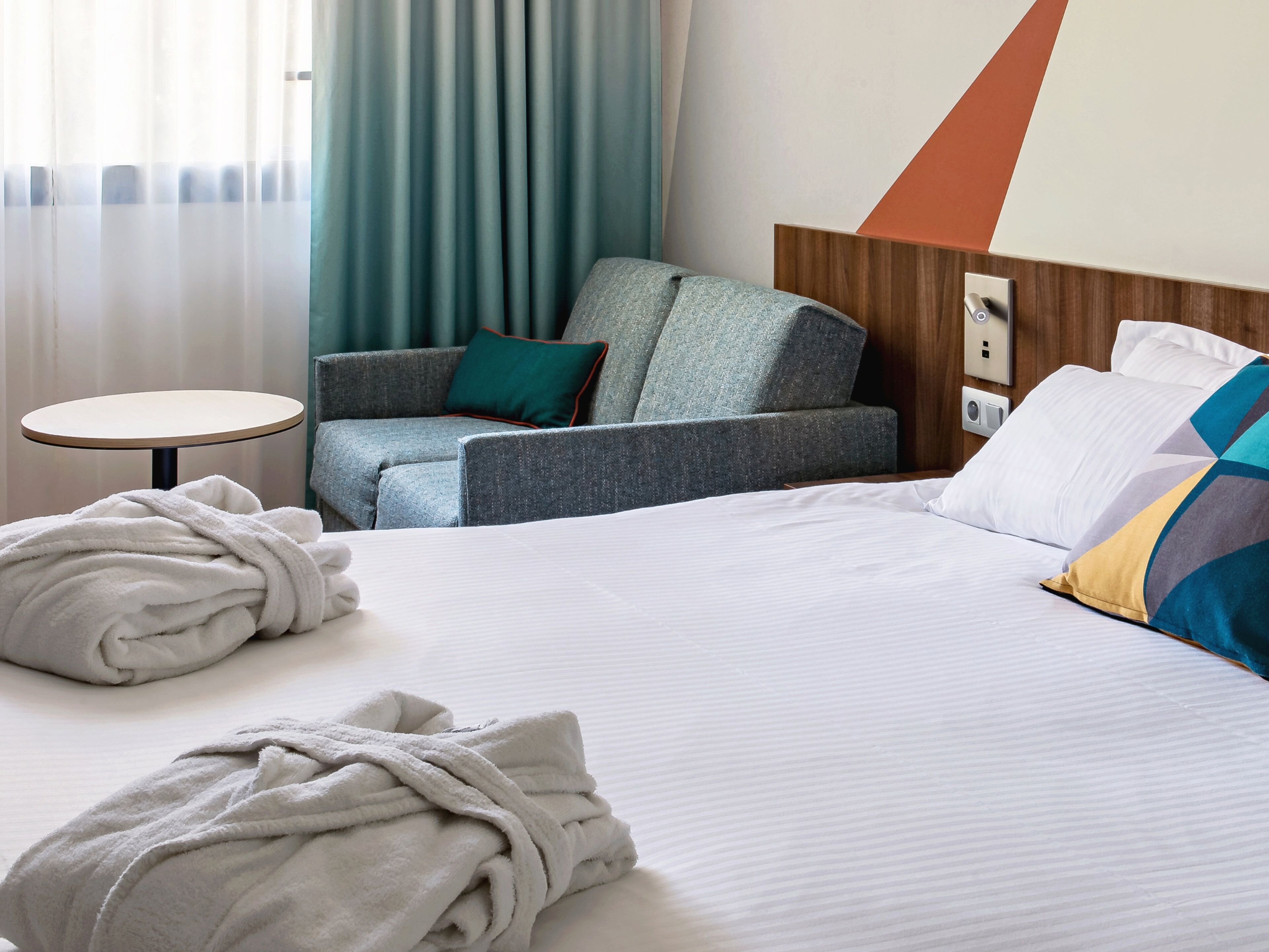 A hotel room with a bed, a round table, a grey armchair with a teal pillow, and a wooden headboard.