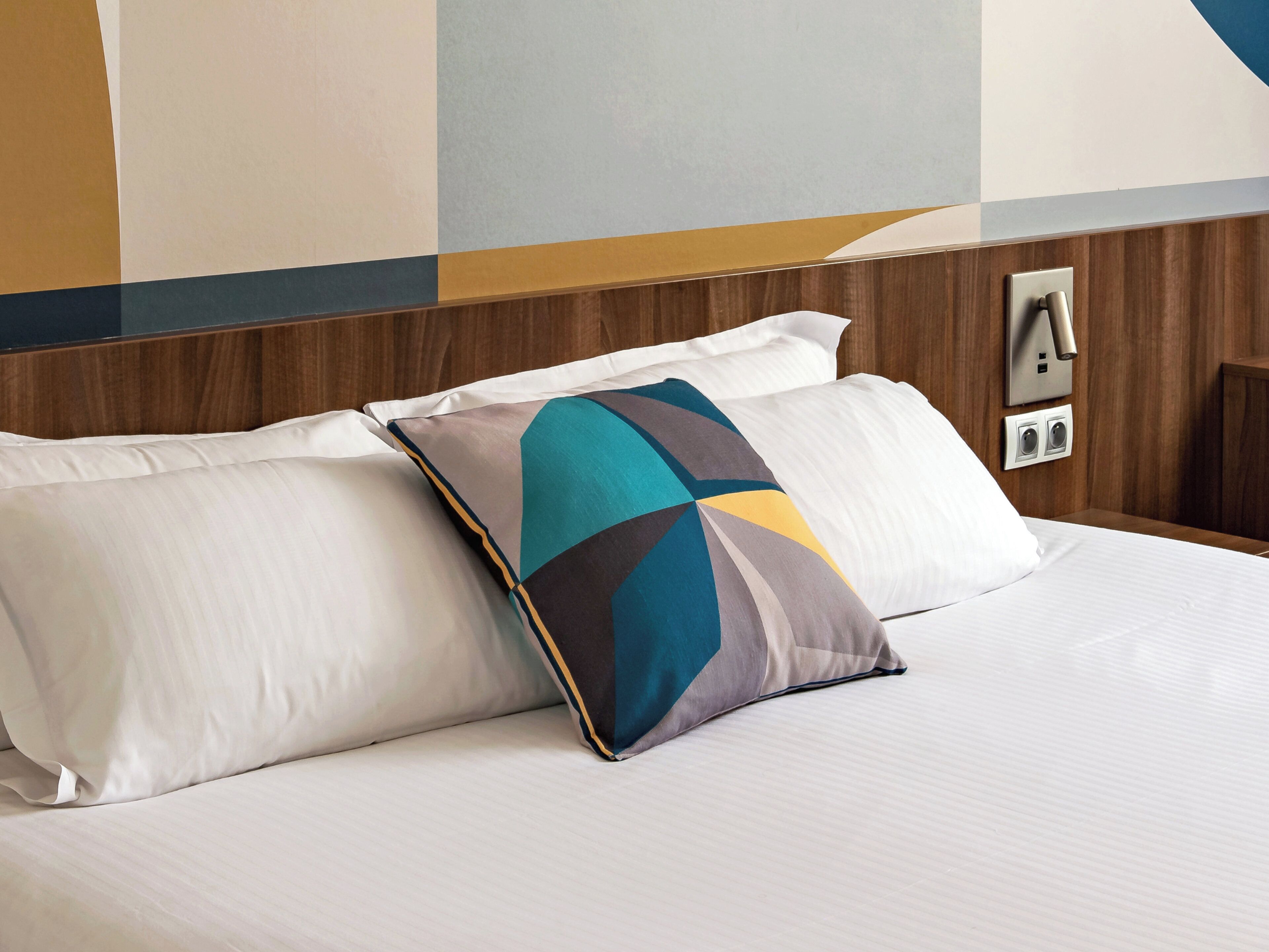 Habitación de hotel con cama, almohadas, cabecero de madera y lámpara de pared.