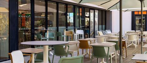 An outdoor seating area with tables and chairs, a large umbrella, and a building with large windows in the background.