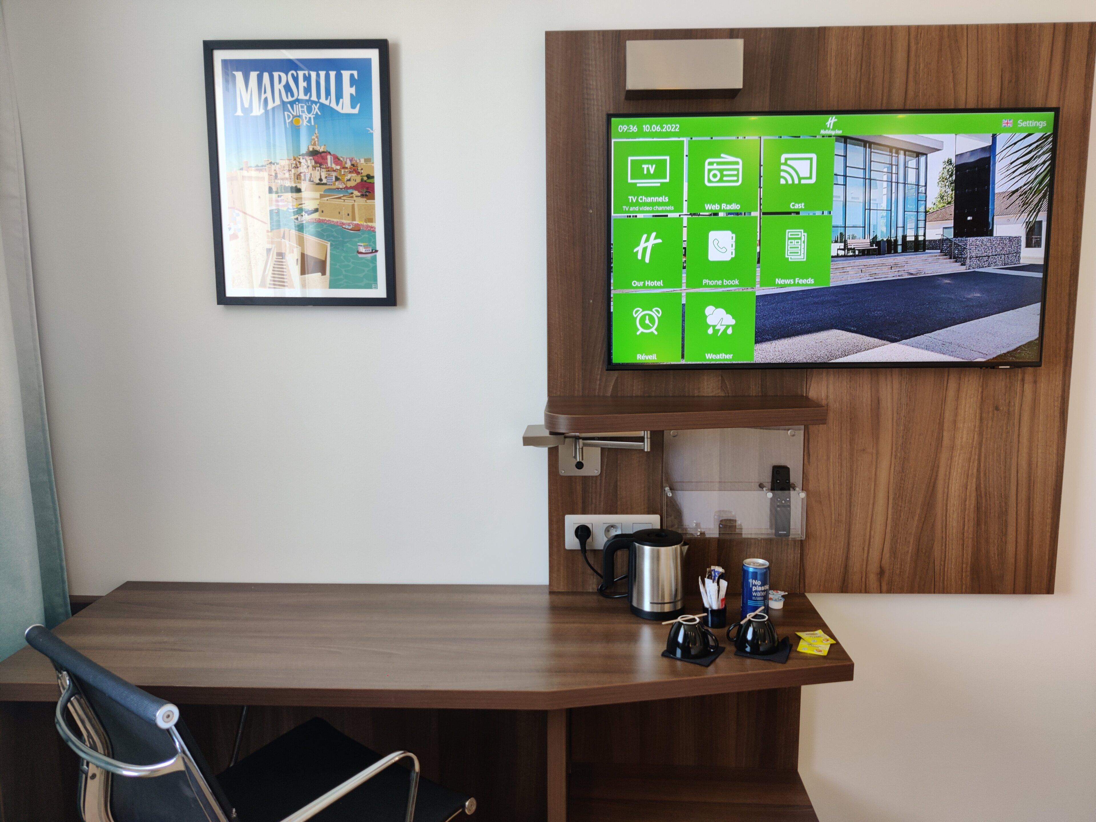 A hotel room with a wooden desk, a framed picture of Marseille, and a digital screen displaying hotel amenities.