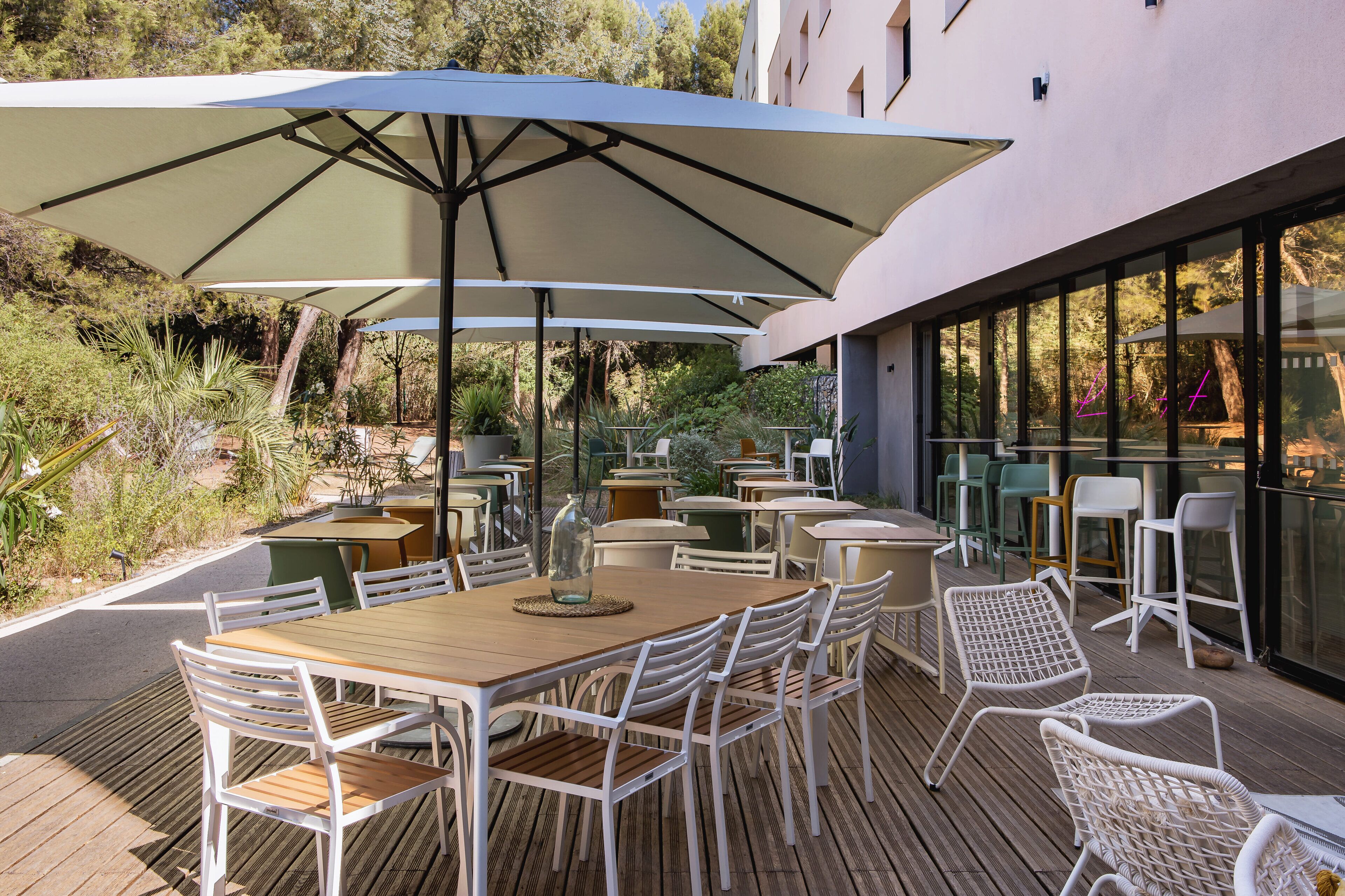 An outdoor patio with wooden tables and white chairs, shaded by large umbrellas, overlooking a garden.