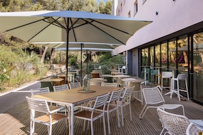 An outdoor patio with wooden tables and white chairs, shaded by large umbrellas, overlooking a garden. - Holiday Inn Marseille Airport by IHG (Vitrolles)