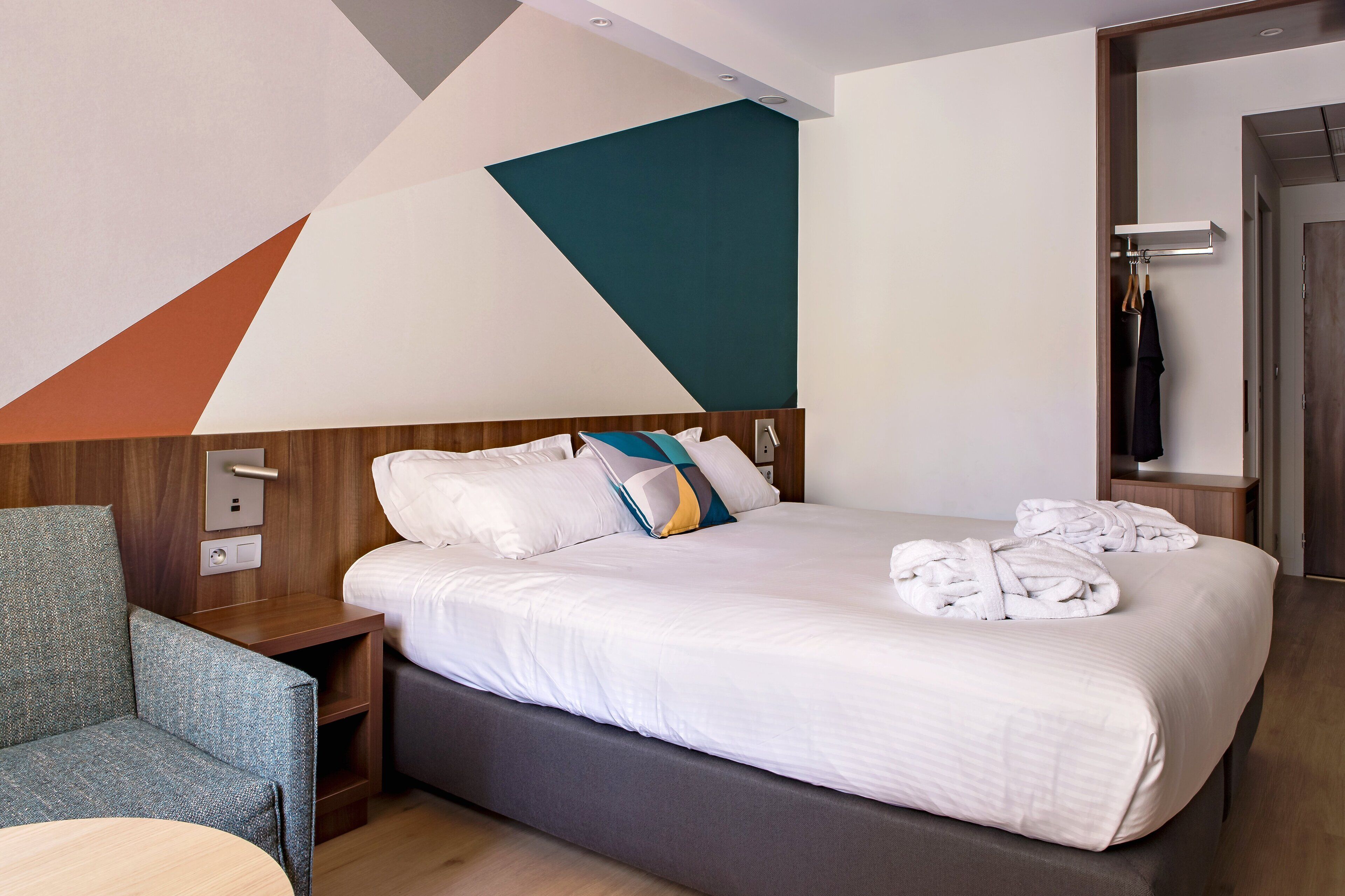 Una habitación de hotel moderna con una cama grande, un cabecero de madera, un sofá gris y una mesita de noche de madera.