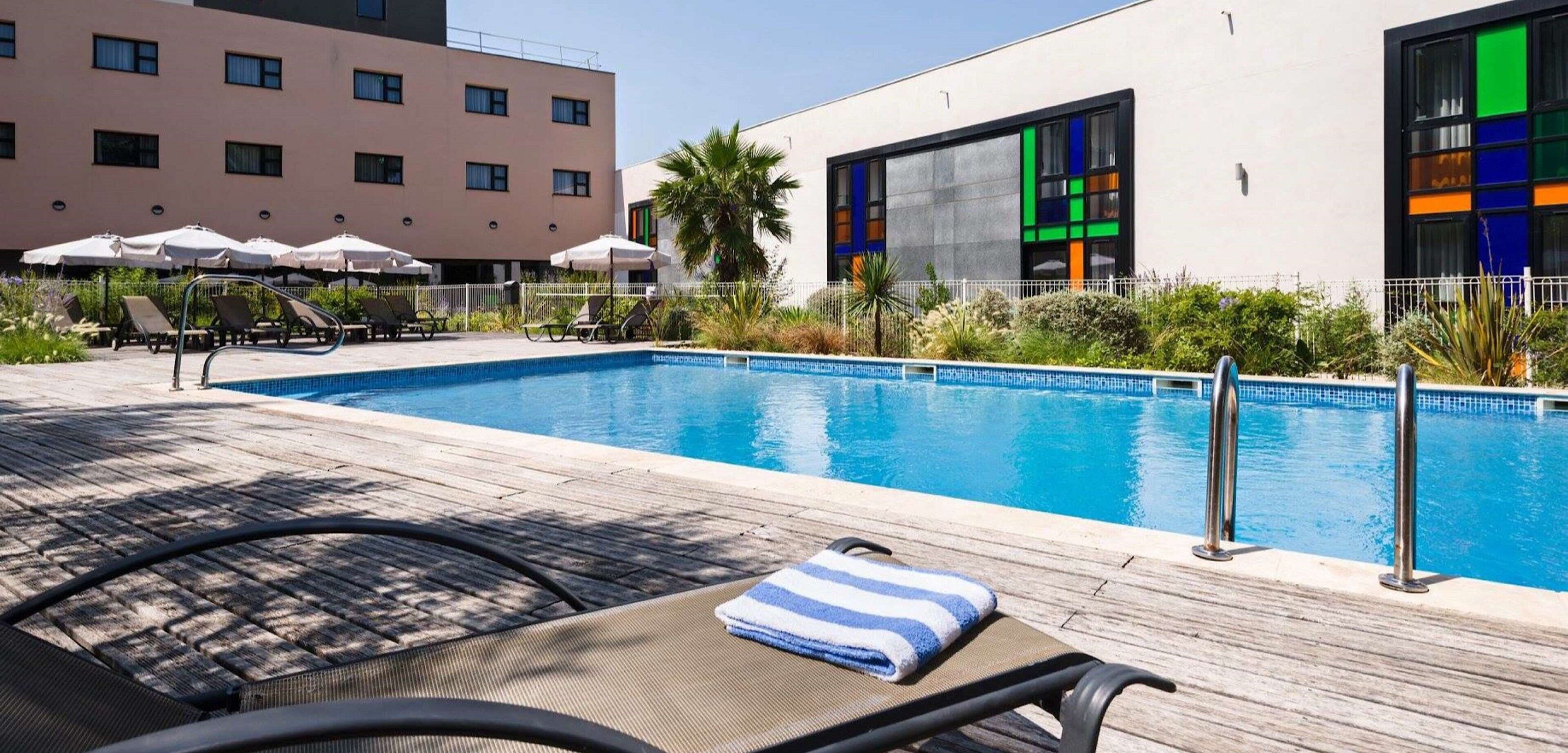 A swimming pool with lounge chairs and umbrellas, surrounded by buildings with large windows and a palm tree.