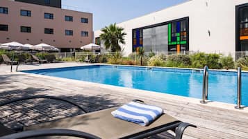 A swimming pool with lounge chairs and umbrellas, surrounded by buildings with large windows and a palm tree.
