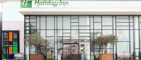 Die Fassade eines Holiday Inn Hotels mit großen Fenstern, einem flachen Dach und zwei Blumentöpfen davor.