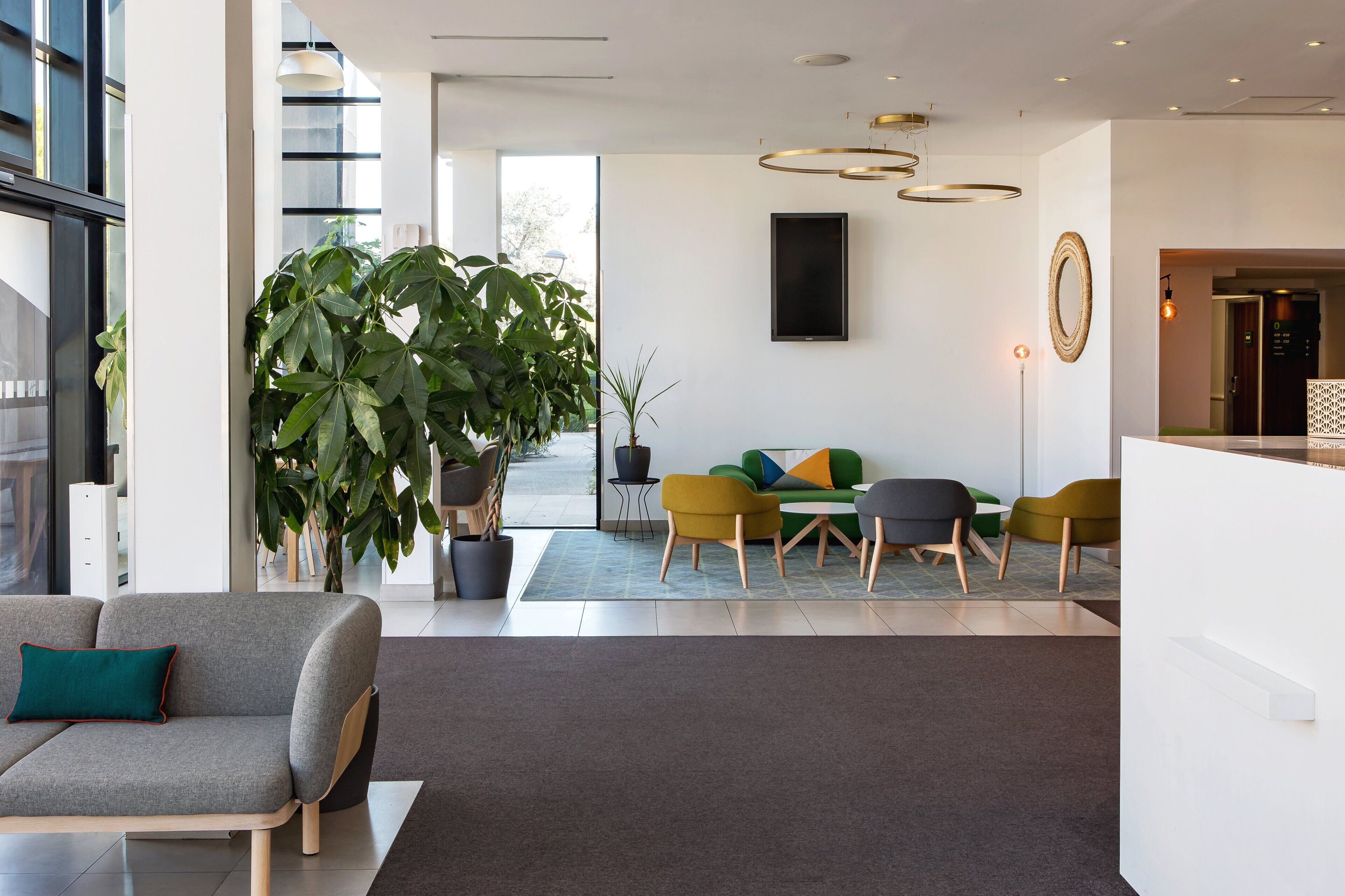 A modern hotel lobby with a sofa, chairs, a large potted plant, and a flat-screen TV.
