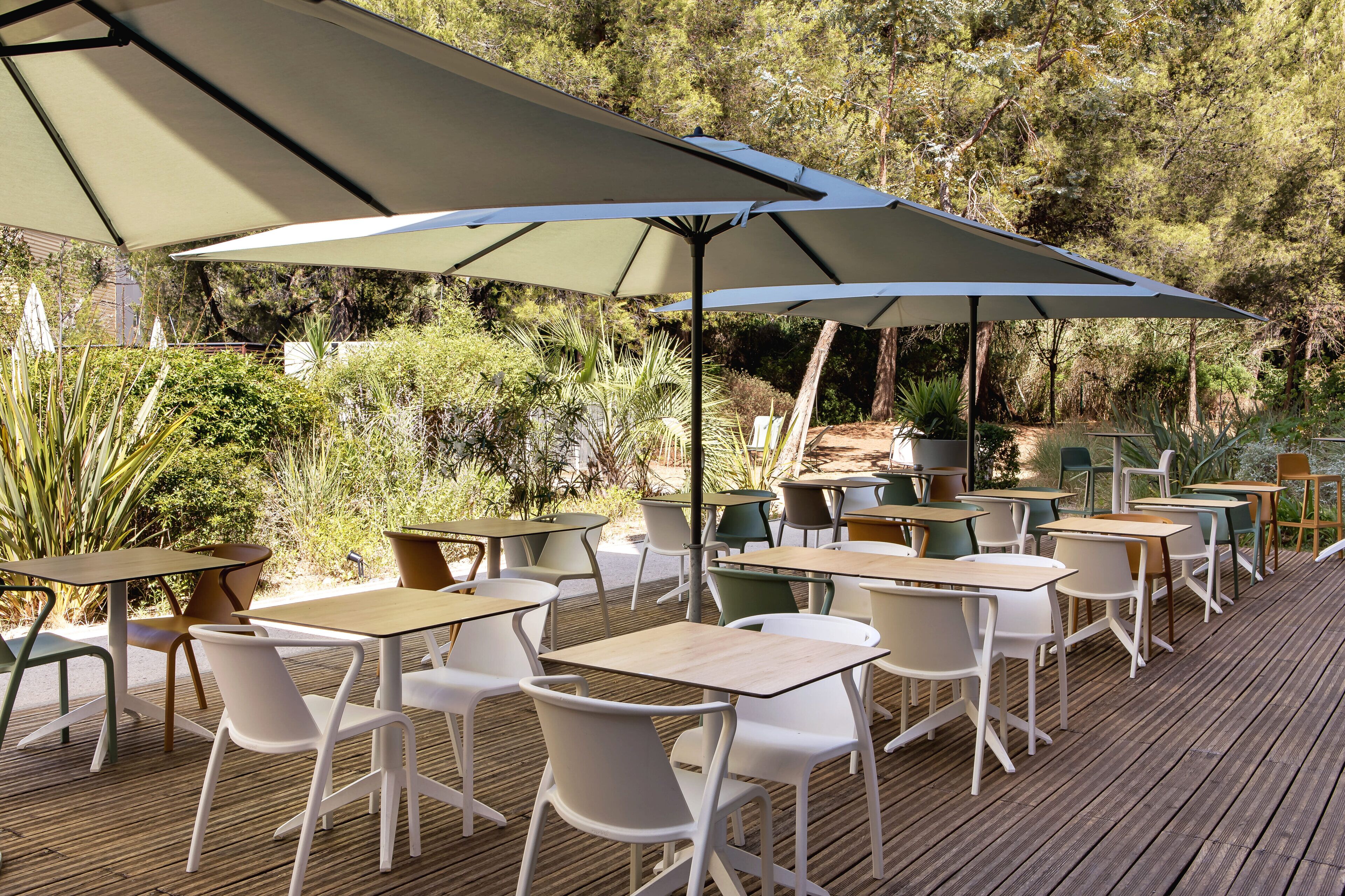 Un espace extérieur aménagé avec des tables et des chaises, à l’ombre de grands parasols, et entouré de verdure.