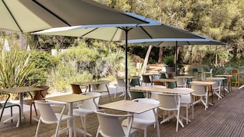 Un espace extérieur aménagé avec des tables et des chaises, à l’ombre de grands parasols, et entouré de verdure.