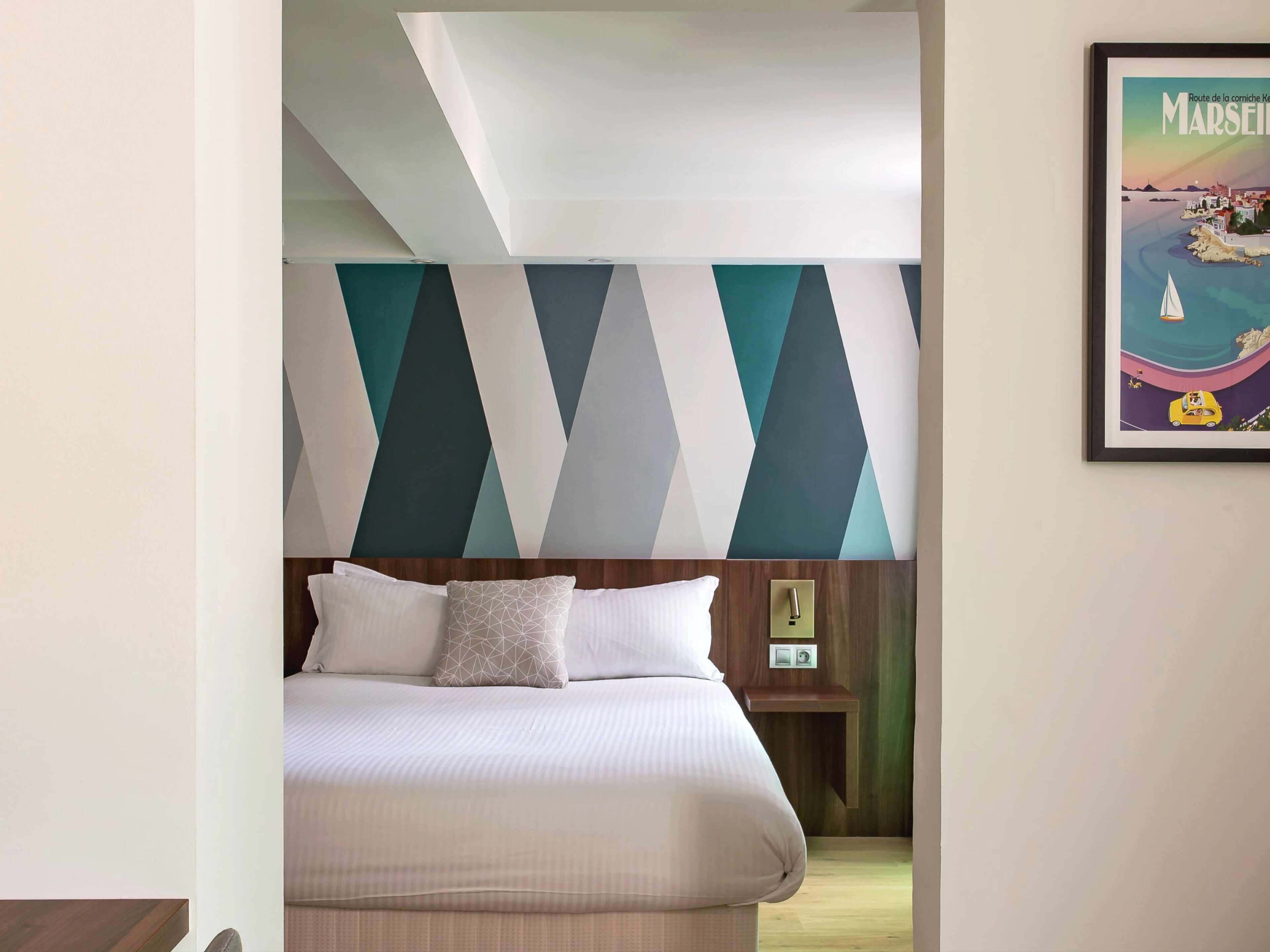 Habitación de hotel con una cama, pared con un diseño geométrico, un cuadro de una escena costera y una mesita de noche con una lámpara.
