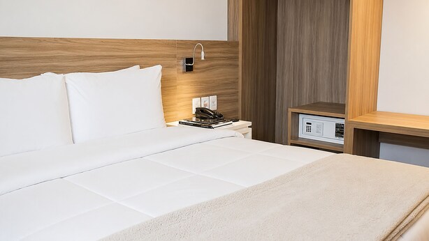 Ein Hotelzimmer mit einem hölzernen Kopfteil, einer weißen Bettdecke, einem Nachttisch mit Telefon und Büchern sowie einer Bedienkonsole.