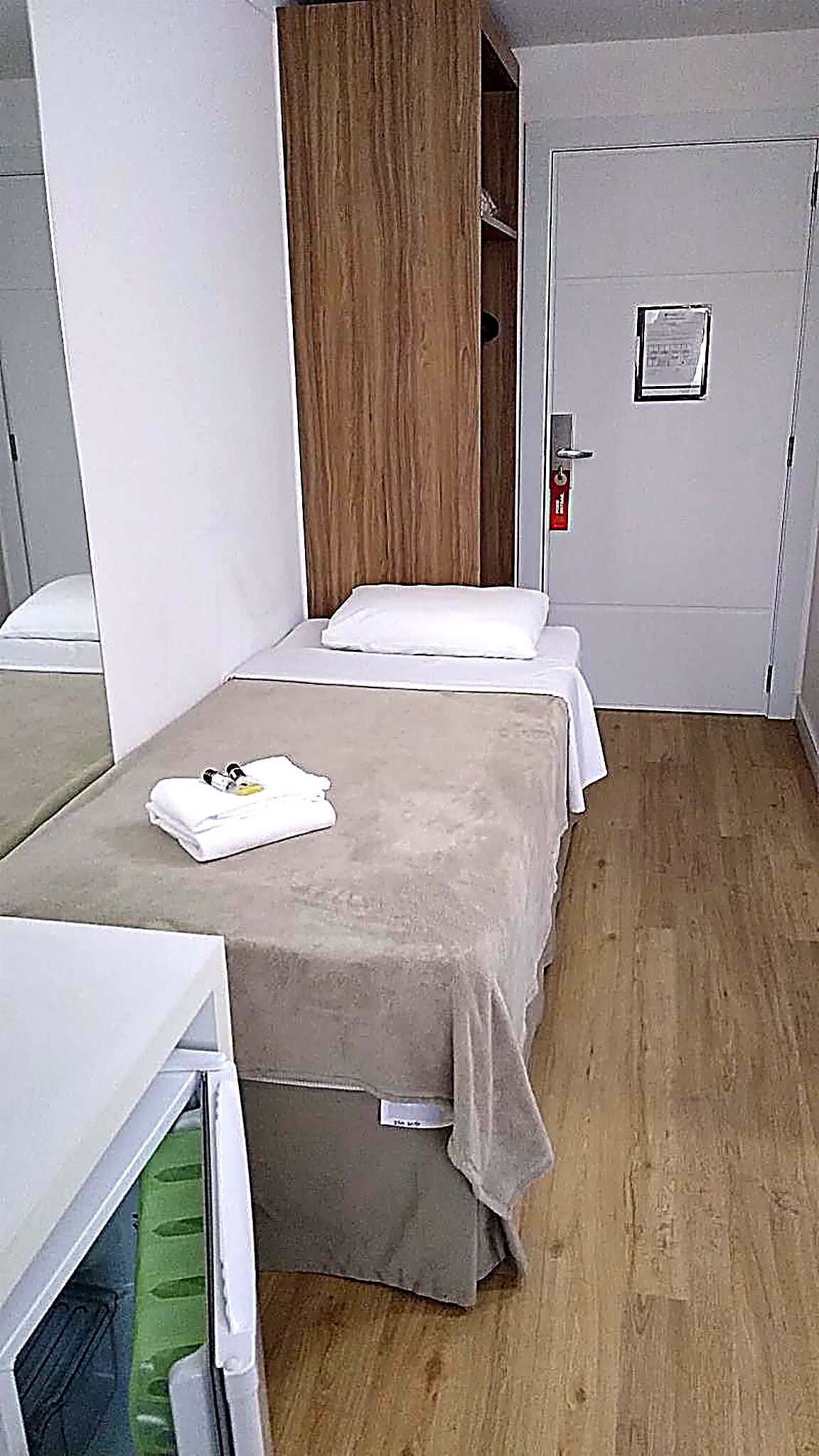 Quarto pequeno com uma cama de solteiro, guarda-roupa de madeira, mesa de cabeceira e uma porta com detector de incêndio.