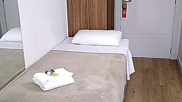 Quarto pequeno com uma cama de solteiro, guarda-roupa de madeira, mesa de cabeceira e uma porta com detector de incêndio.