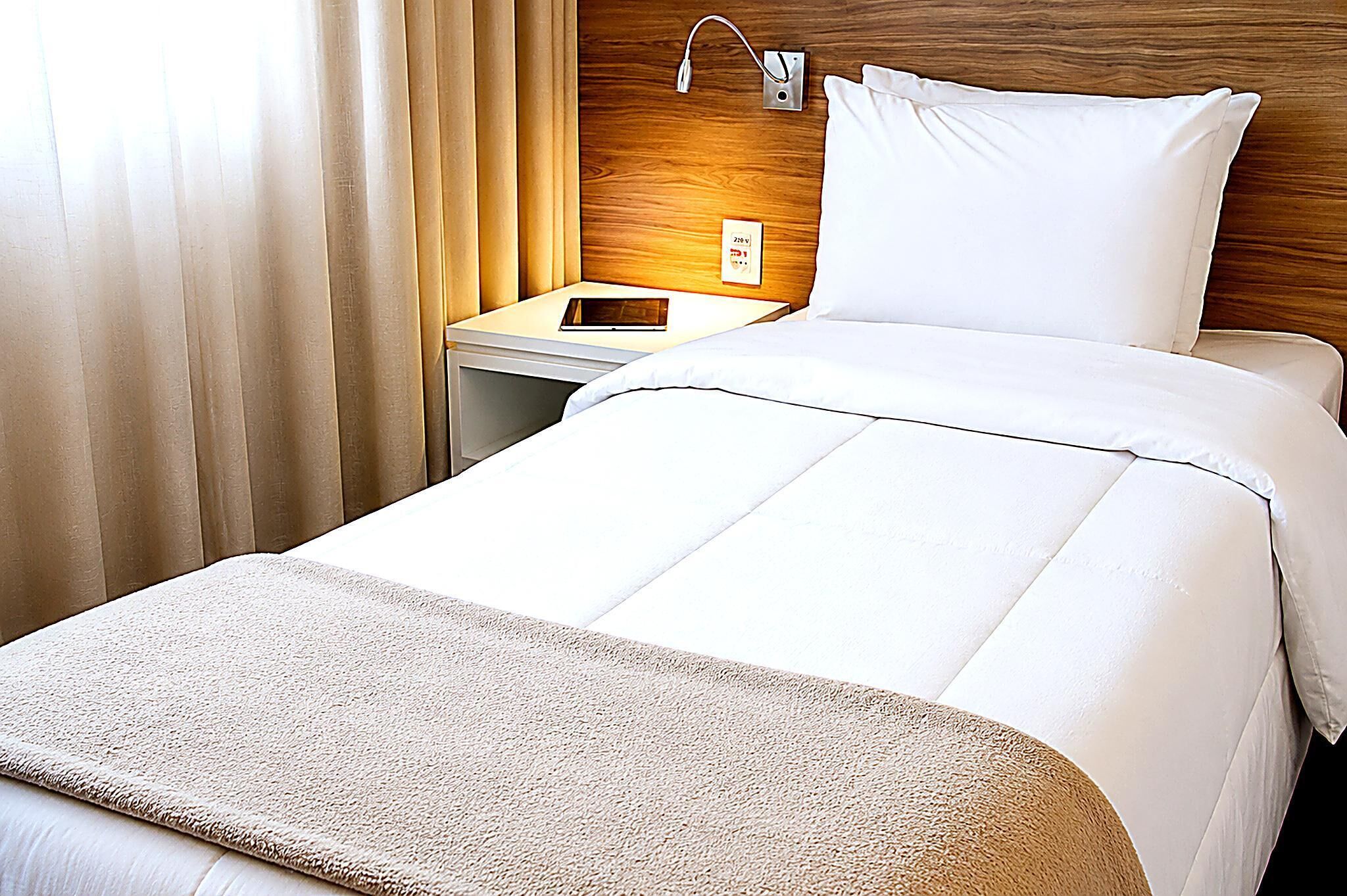 A hotel room with a bed, a bedside table, a nightstand, a lamp, and a power outlet.