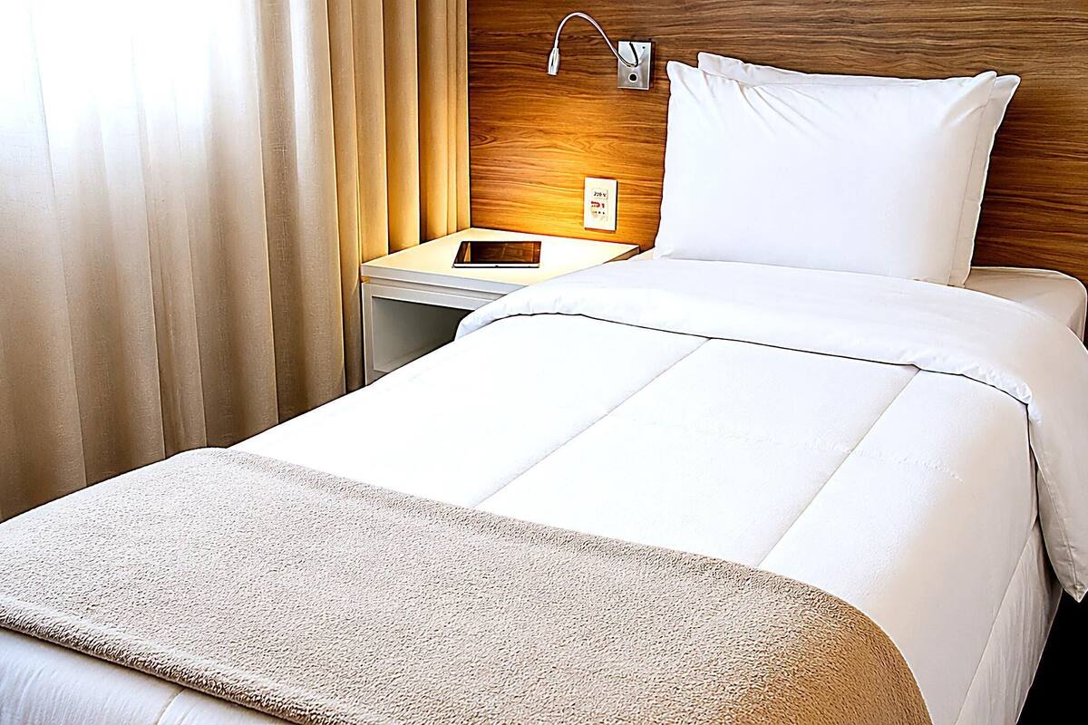 Una habitación de hotel con cama, mesita de noche, lámpara y toma de corriente.
